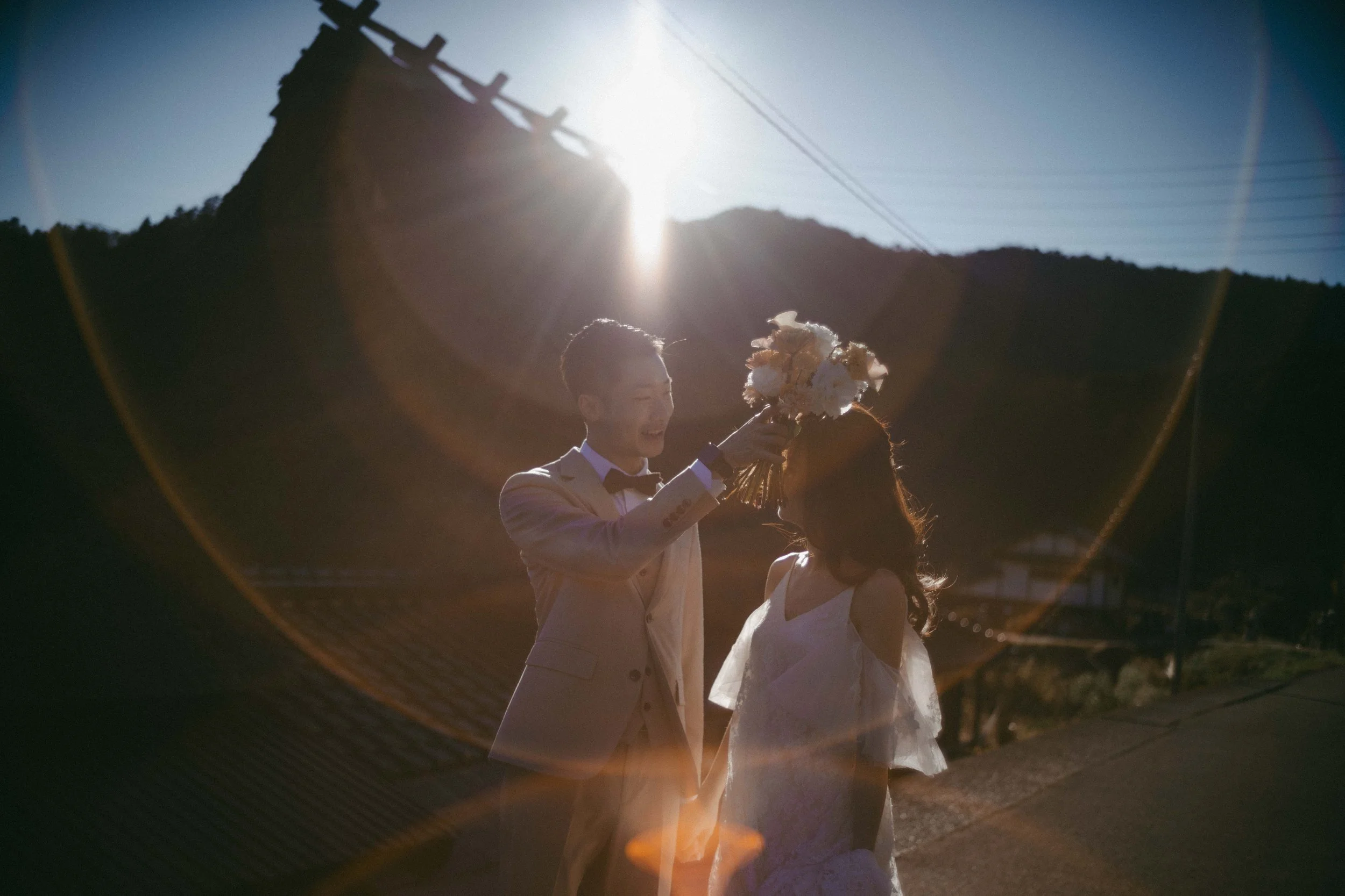 A man in a beige suit and a woman in a white dress share a moment outdoors, with the man holding a bouquet of flowers above the woman's head, backlit by the sun creating a lens flare. Mountains and power lines are in the background.
