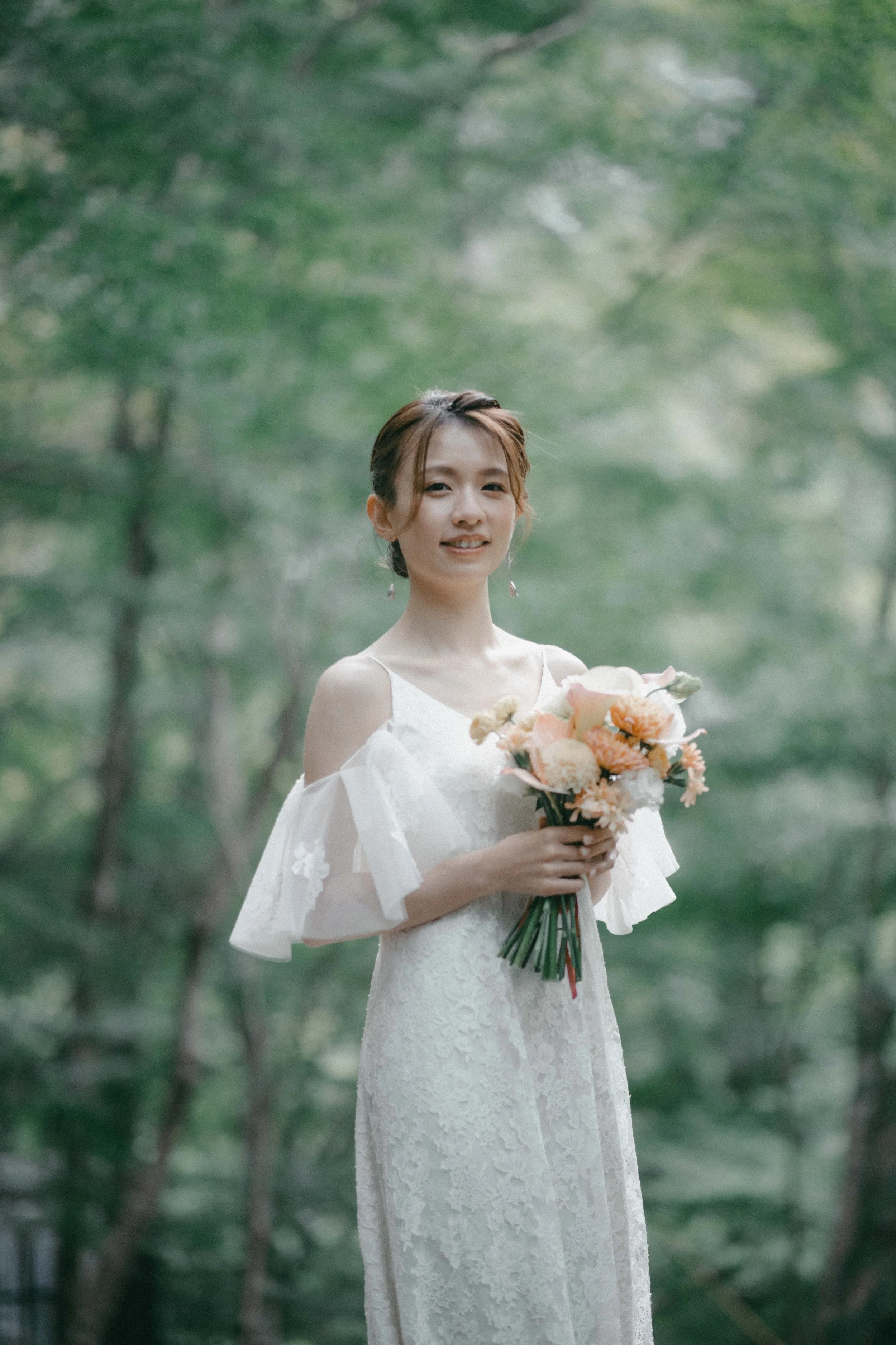 A woman in a white lace wedding dress holding a bouquet of light-colored flowers in a green, outdoor setting.