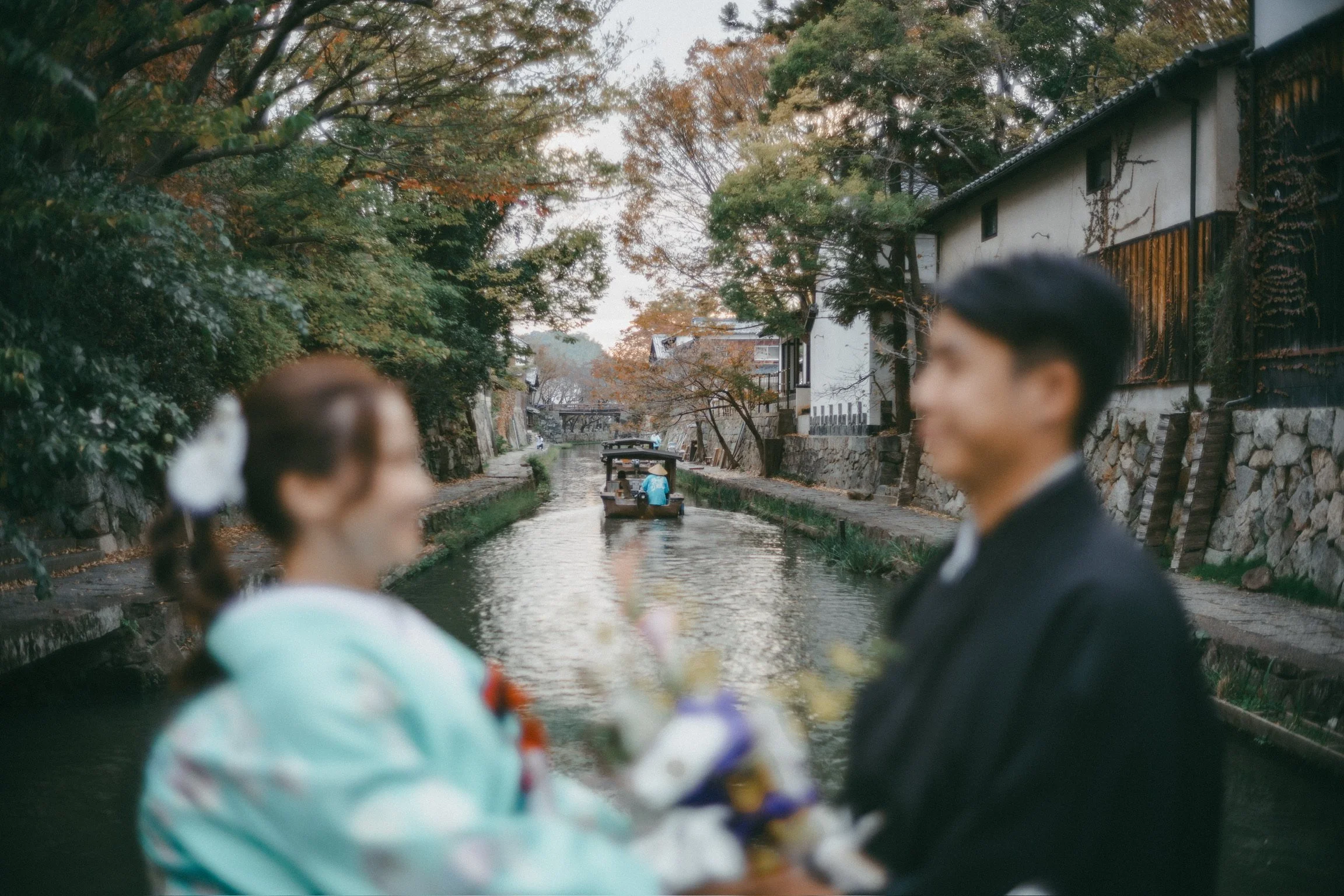 A couple dressed in traditional Japanese wedding attire stands on a canal side in a scenic, historic area with wooden buildings and lush trees. A boat with passengers is visible in the background.