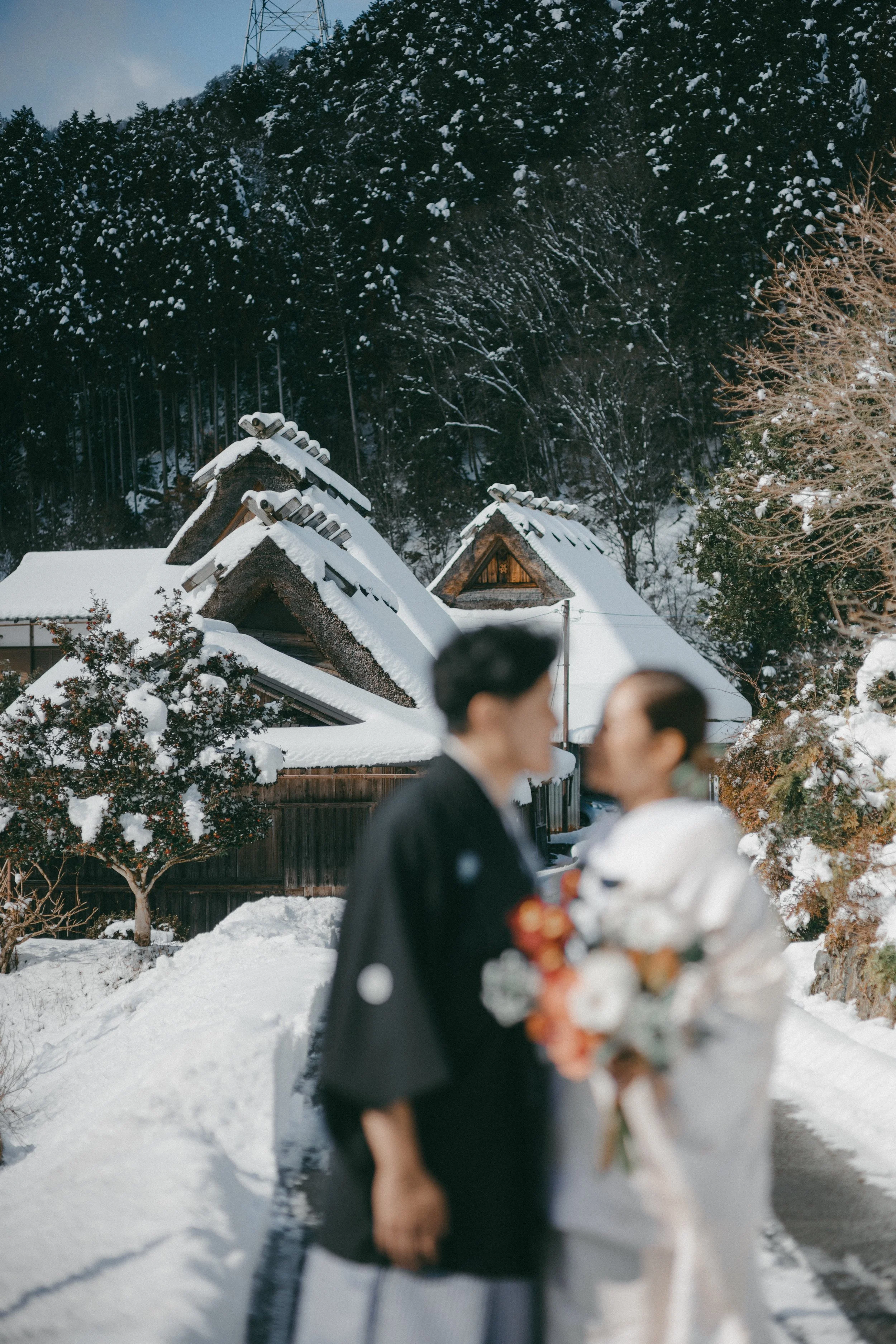 A blurry couple in wedding attire standing outdoors in a snowy landscape with traditional Japanese houses and trees in the background.
