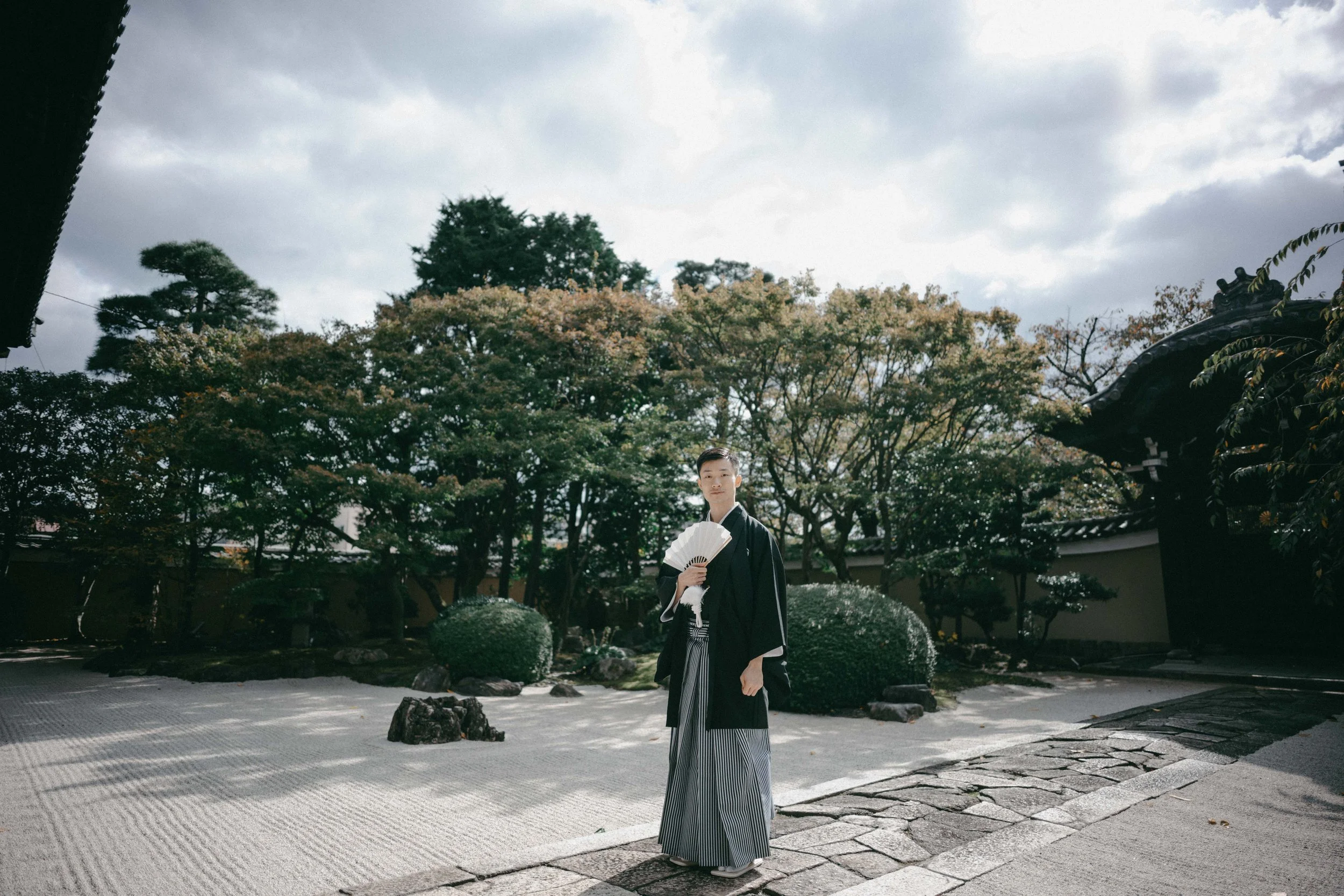 A young man wearing traditional Japanese clothing standing in a Japanese garden, holding a folding fan, with trees and a traditional roofed structure in the background.