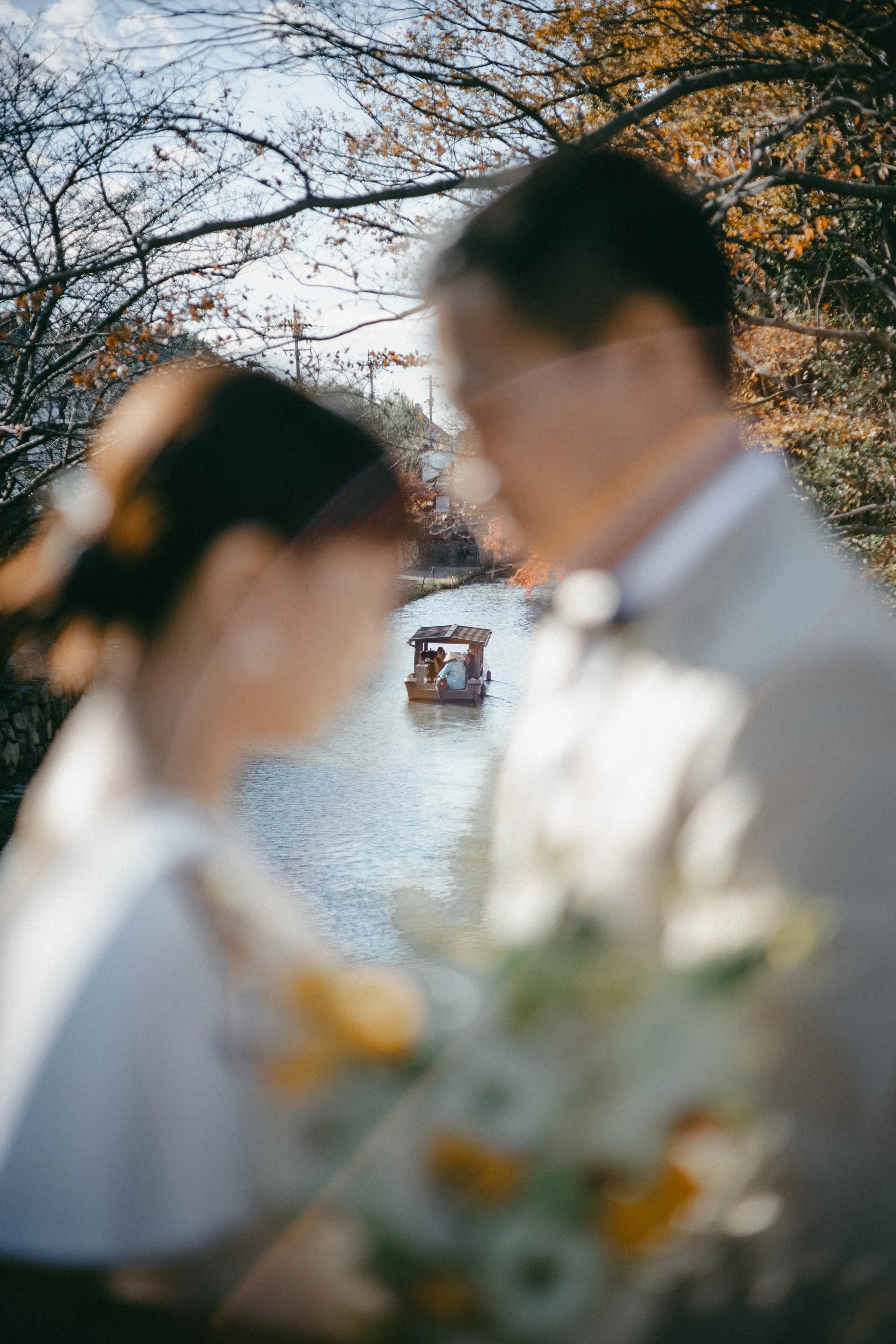 A blurry photograph of a bride and groom outdoors near a river, with a boat floating on the water in the background, during autumn.