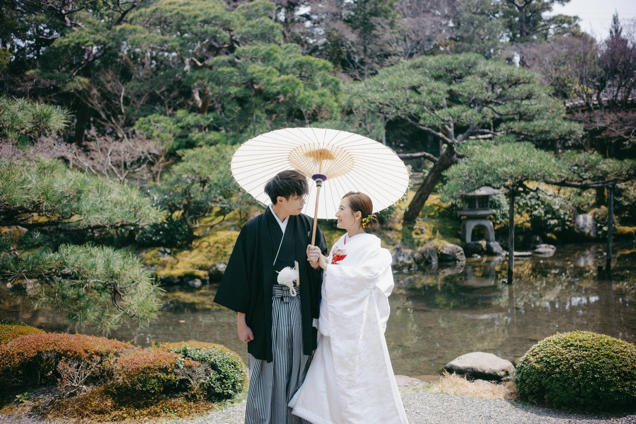 A couple in traditional Japanese wedding attire standing in a garden, holding a white umbrella, with greenery and a pond in the background.