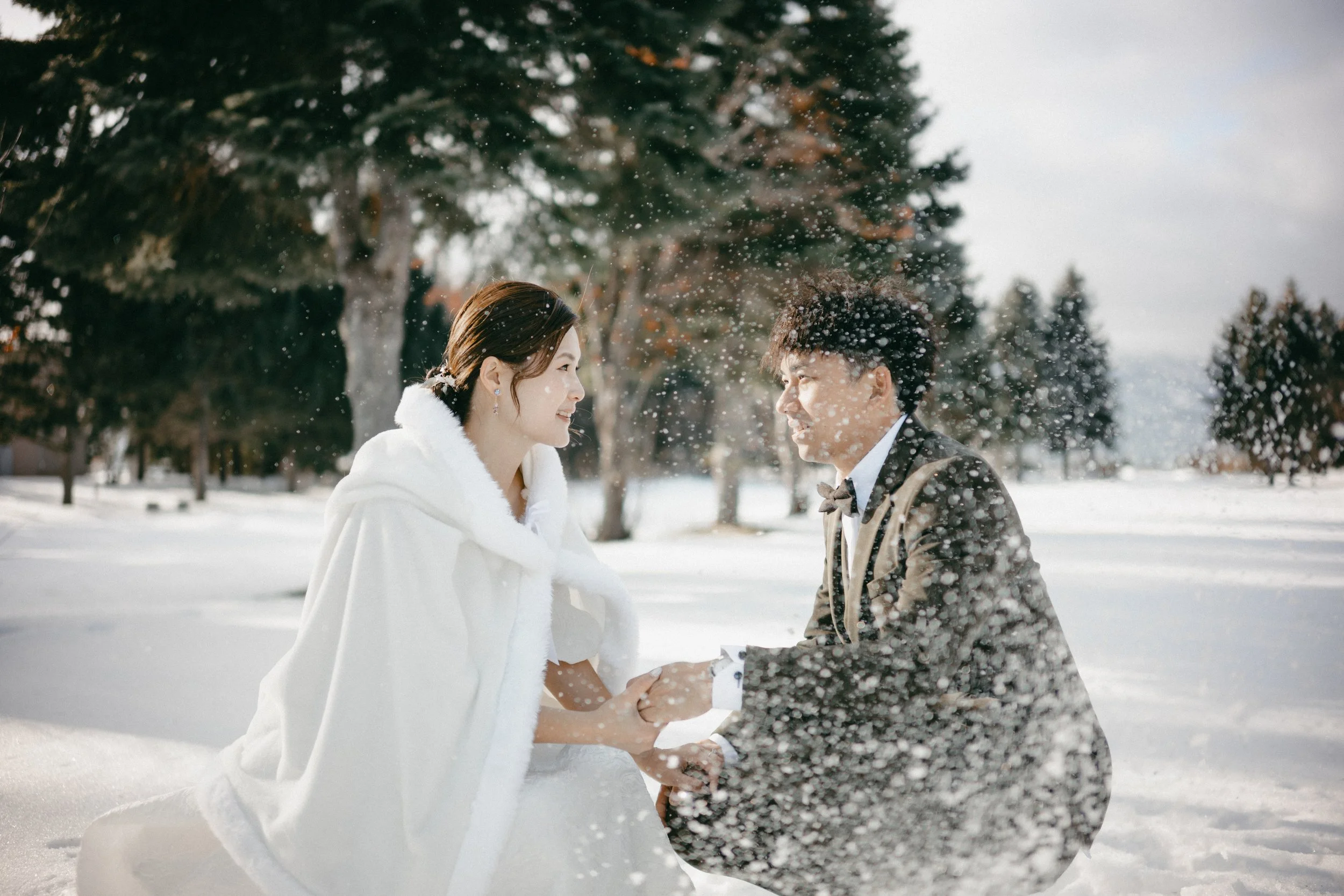 A couple dressed in wedding attire in a snowy outdoor setting, holding hands and smiling at each other amidst falling snow.