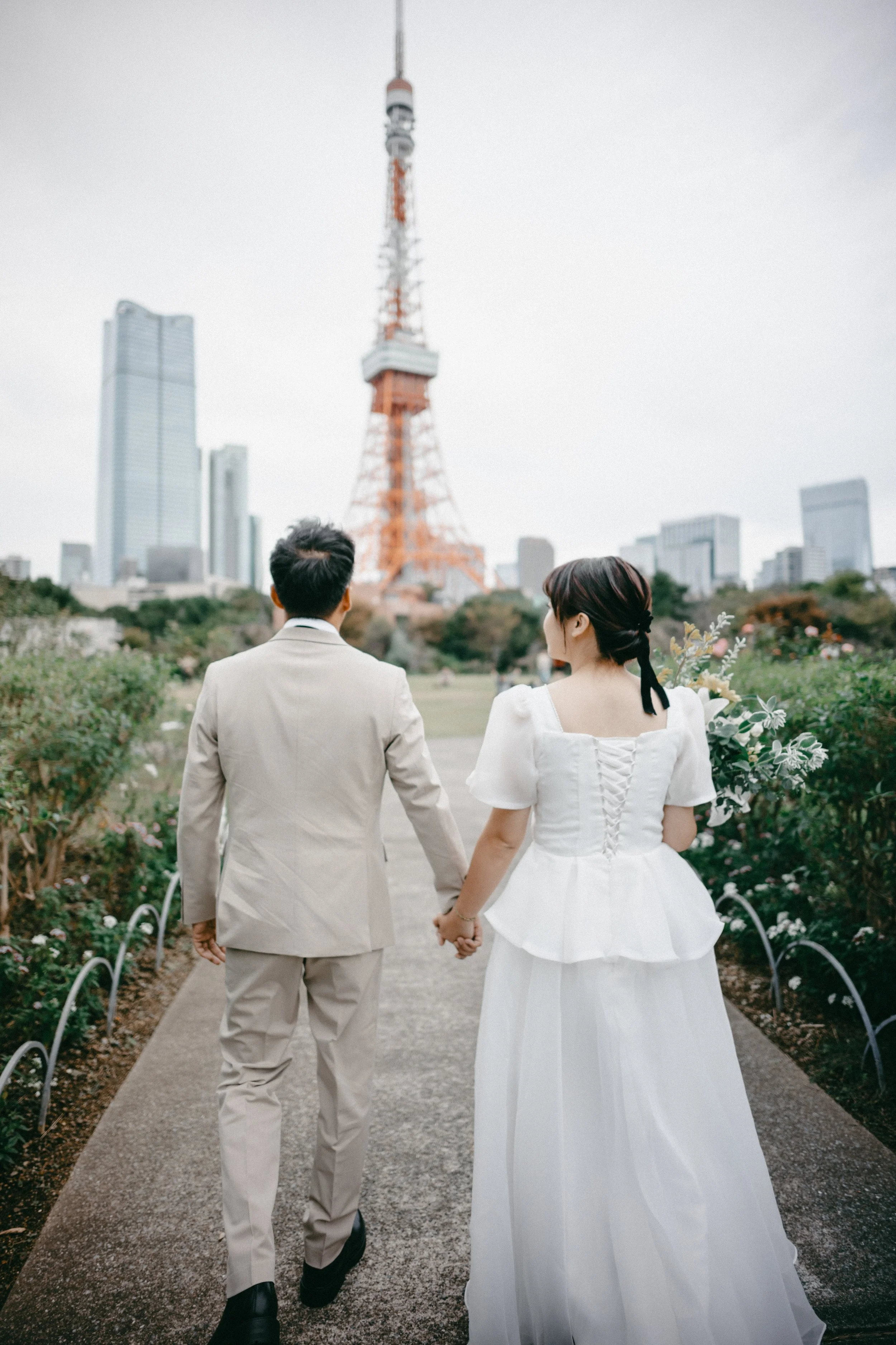 A couple dressed in wedding attire walking hand in hand through a garden with Tokyo Tower in the background.