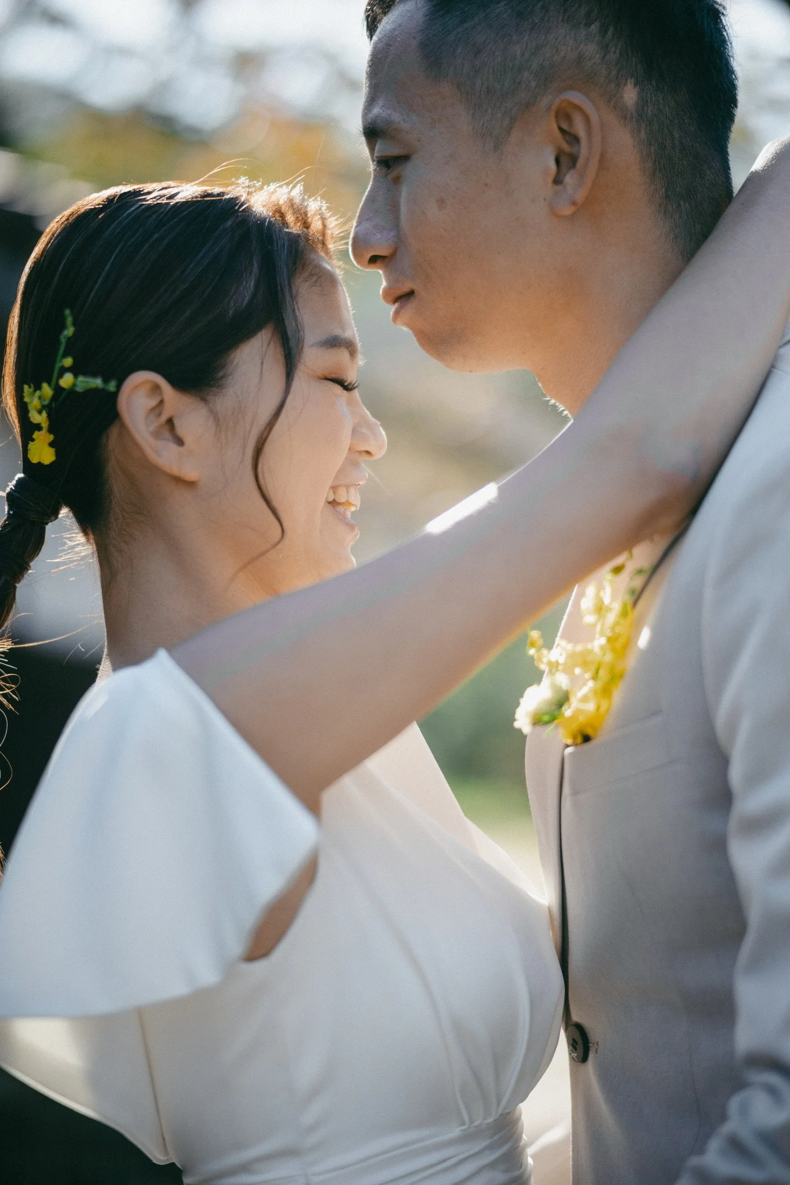 A joyful couple on their wedding day, close together outdoors, smiling, with sunlight and greenery in the background.