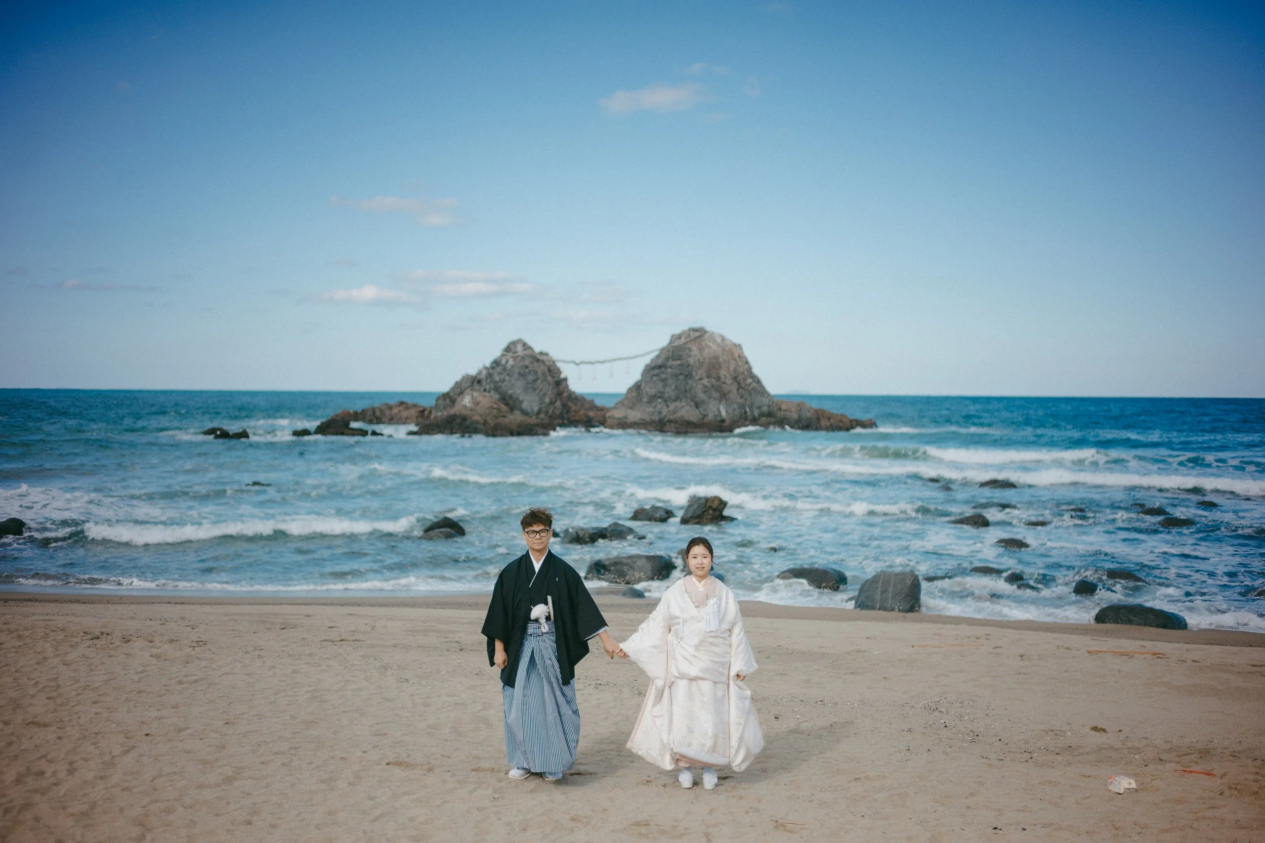 A couple in traditional Japanese wedding attire holding hands on a beach with rocks and ocean in the background.