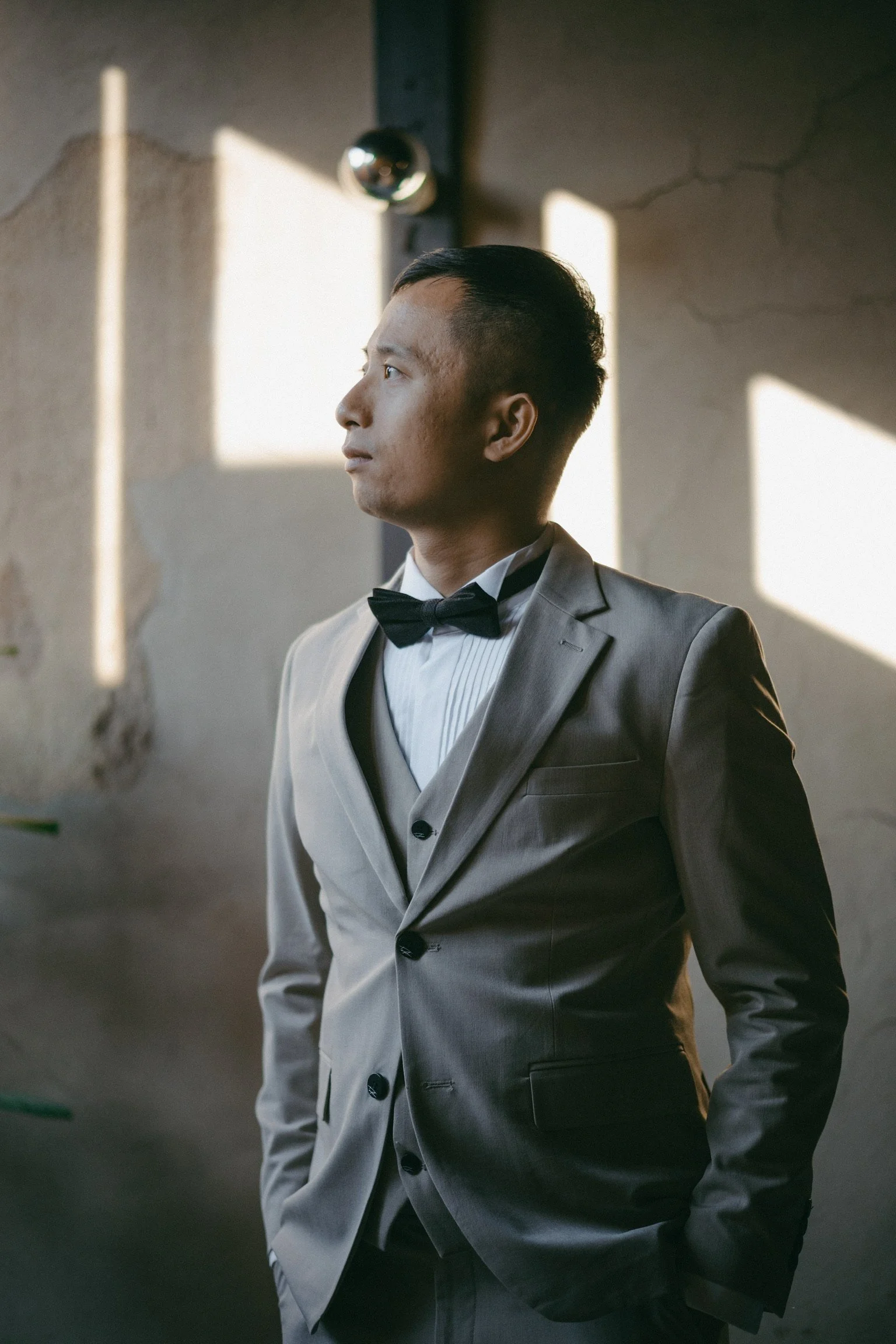 A man dressed in a beige tuxedo with a white shirt and black bow tie stands sideways near a wall, looking contemplatively to his left. Sunlight streams through window openings, casting light and shadow on the wall behind him.