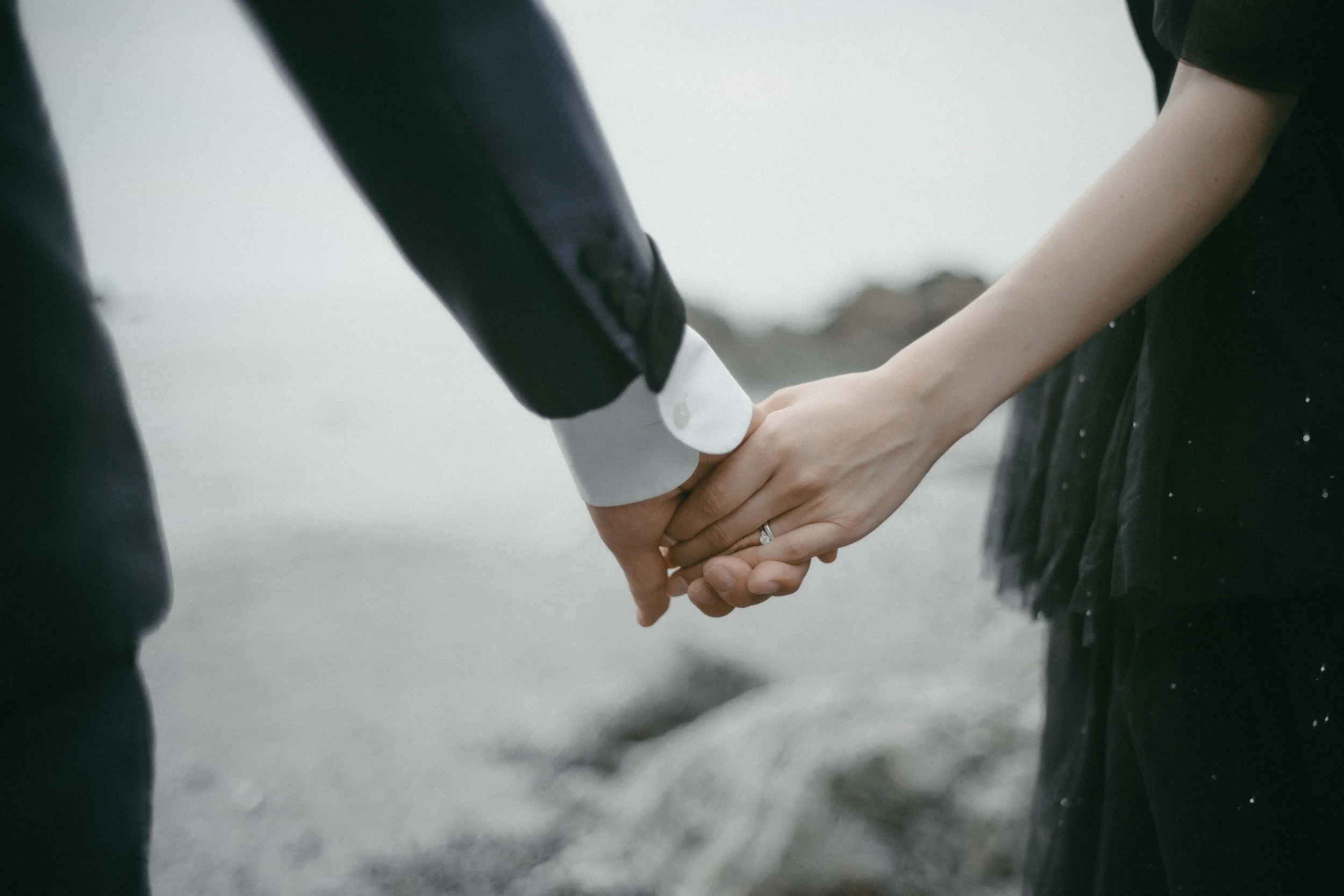 Close-up of a man and woman holding hands outdoors on a beach, wearing formal attire.