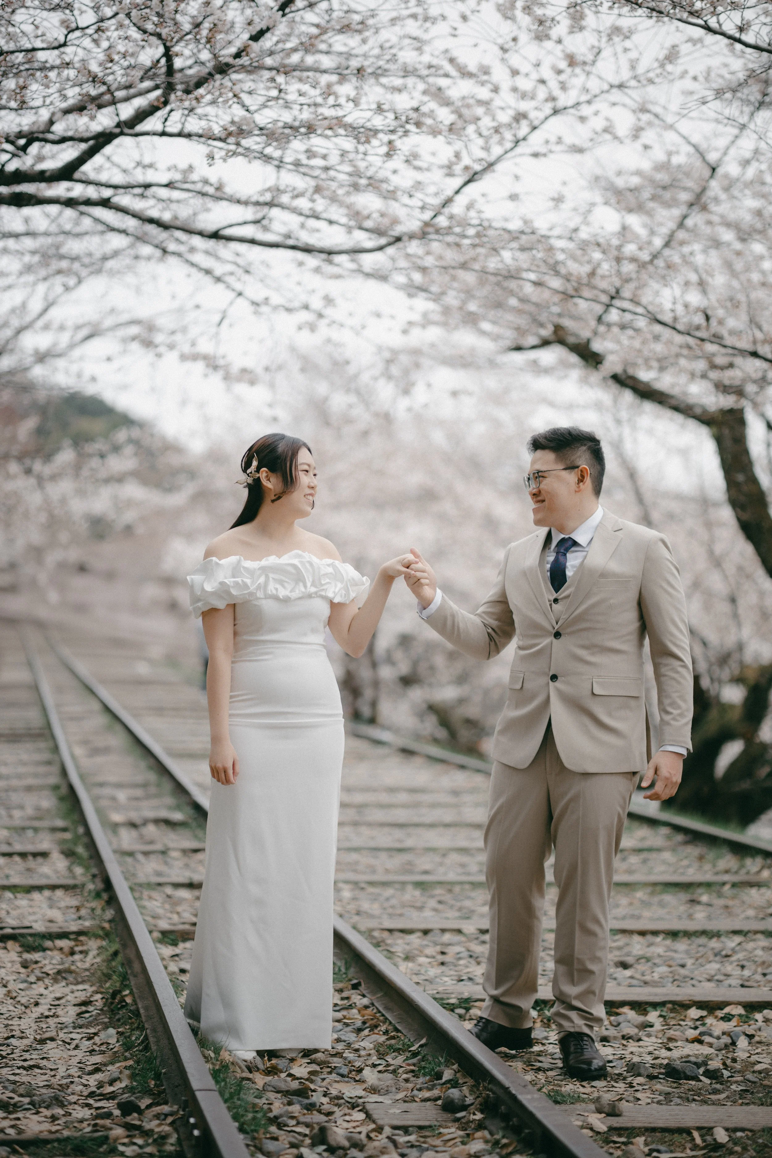 A couple dressed in formal wedding attire holding hands on train tracks under cherry blossom trees.