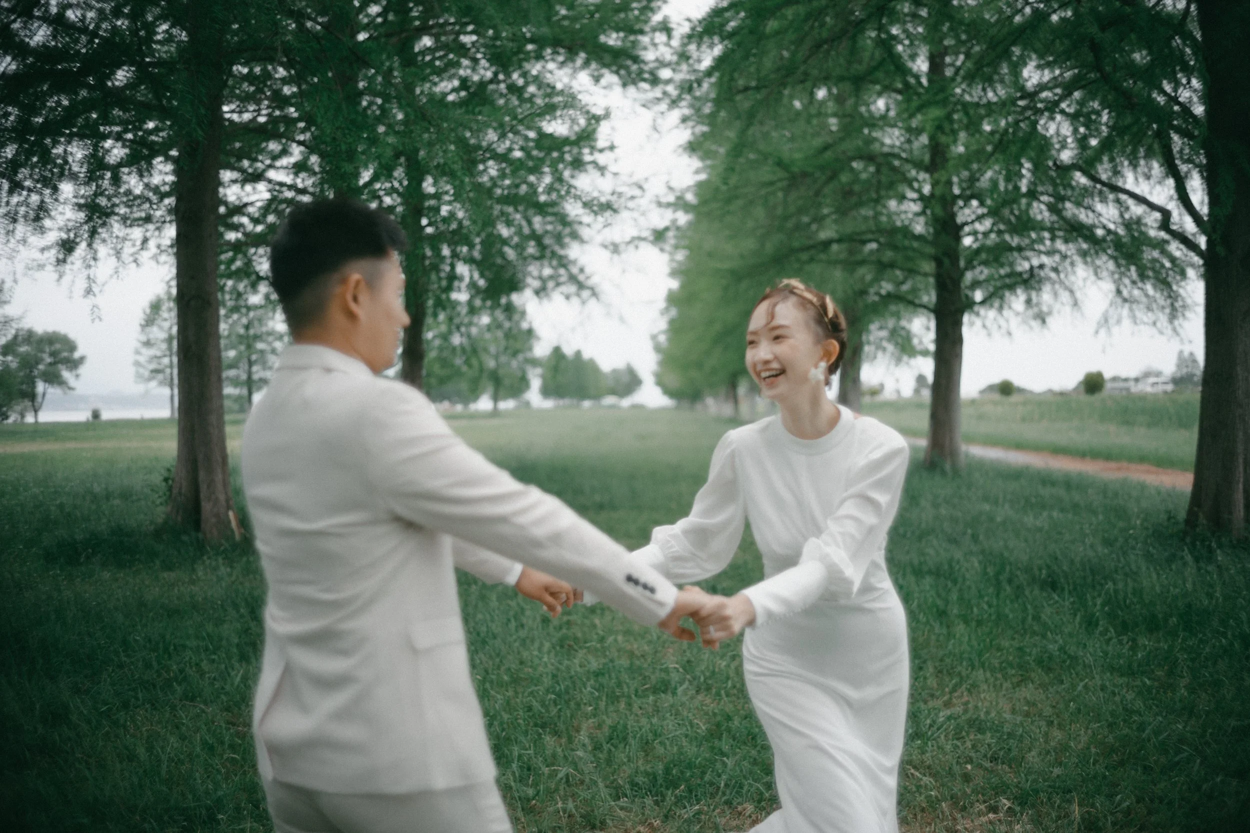 A couple dressed in white holding hands and laughing in a park with trees and grass.