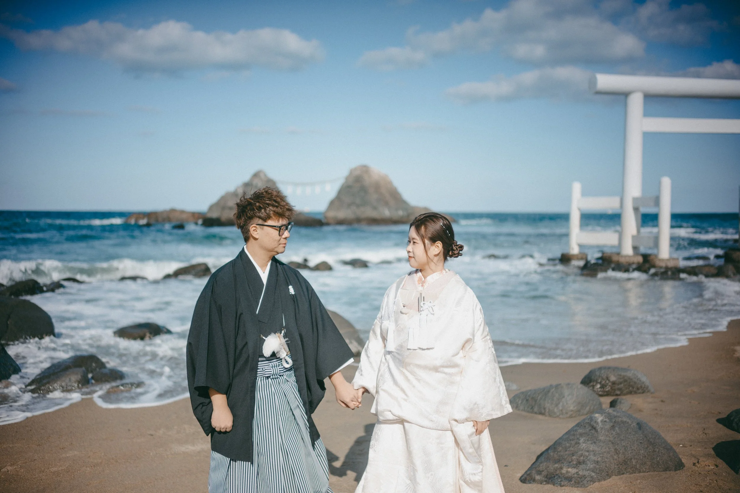 A couple in traditional Japanese wedding attire holding hands on a beach with rocks, ocean waves, and a torii gate in the background.