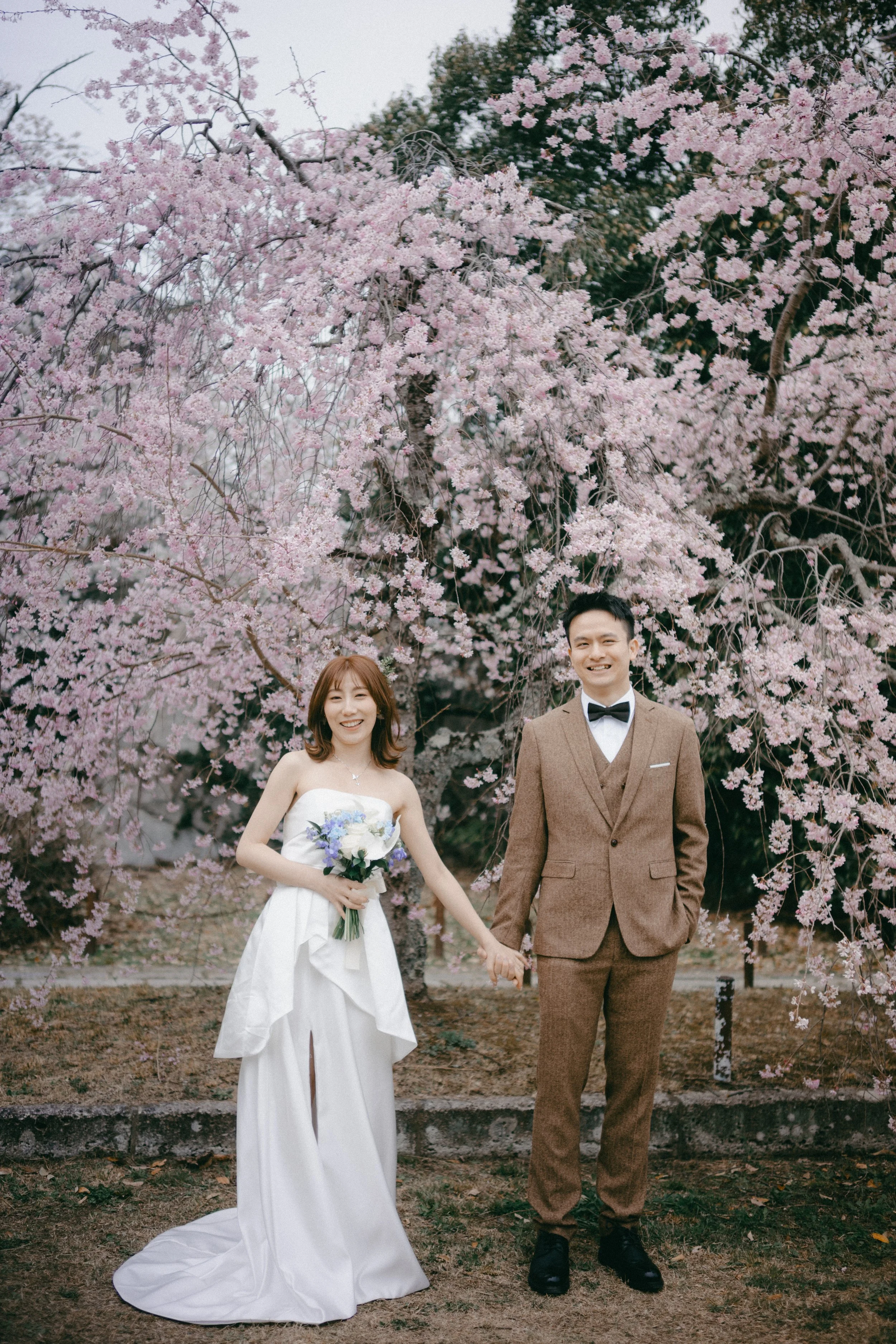 A bride and groom holding hands in front of a blooming cherry blossom tree. The bride is in a white strapless wedding gown holding a bouquet of white and purple flowers, and the groom is in a brown suit with a bow tie.
