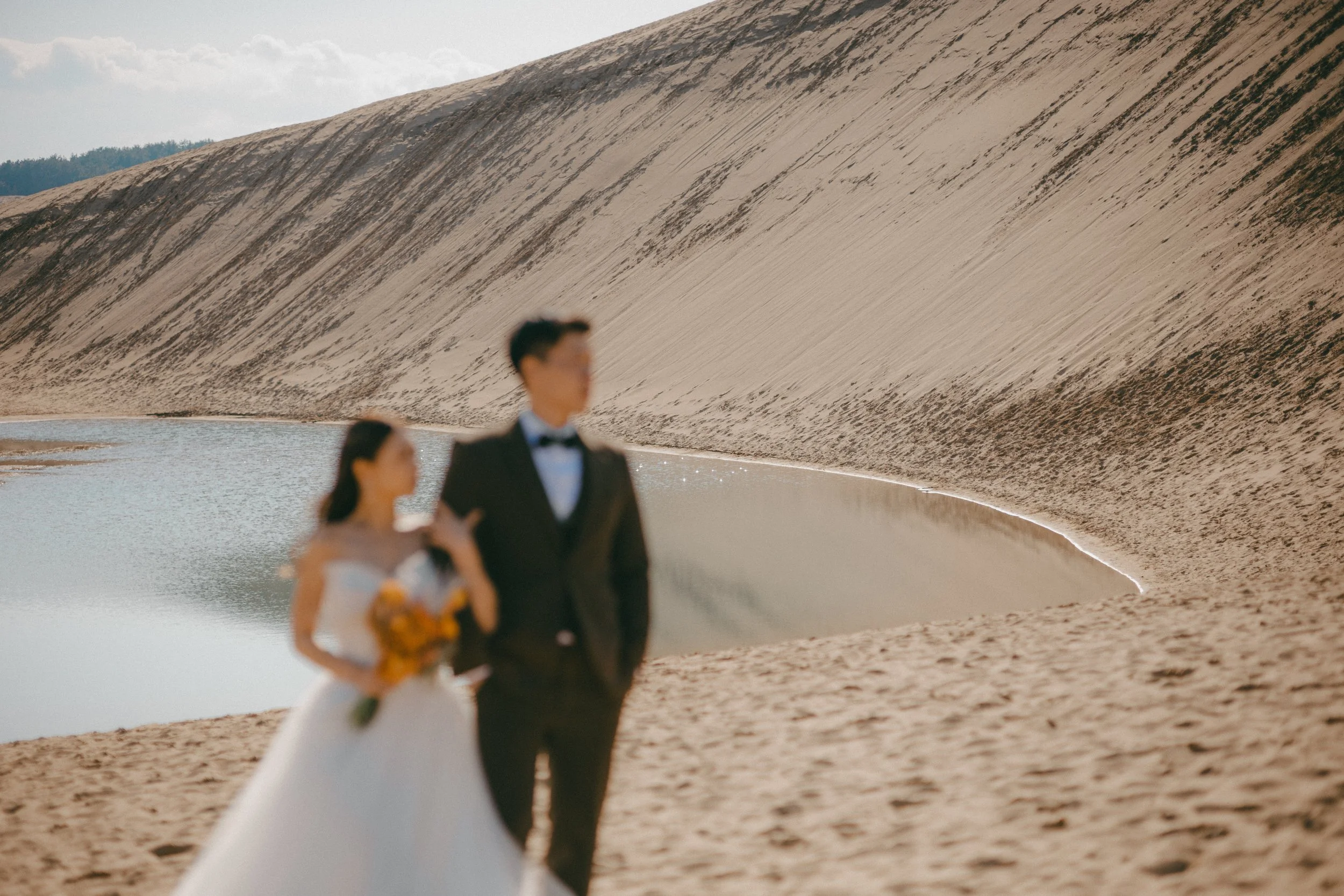 A bride and groom in wedding attire standing on sandy terrain near a small lake with a large sand dune in the background.