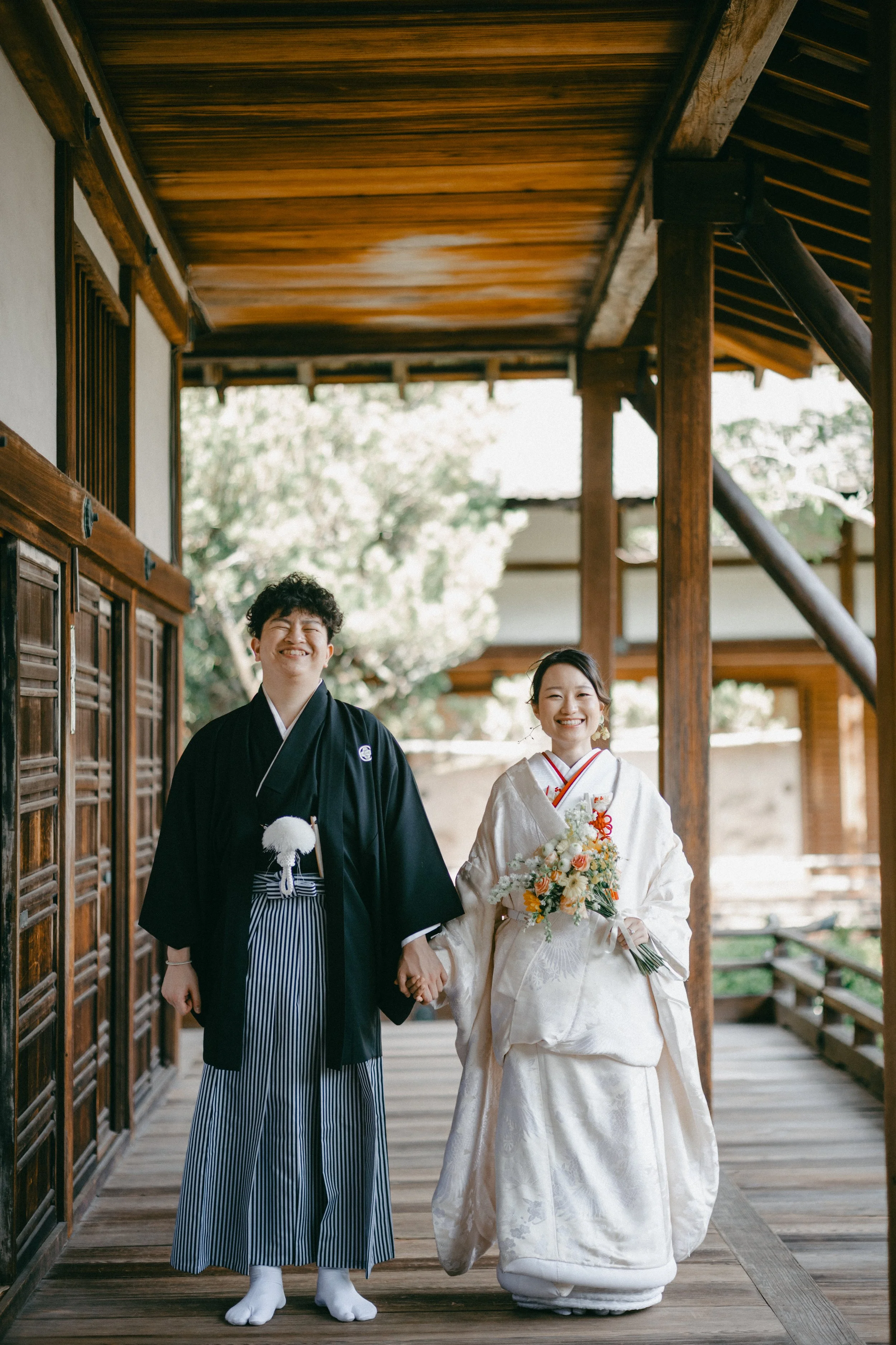 A wedding couple in traditional Japanese attire holding hands and smiling on a wooden porch.