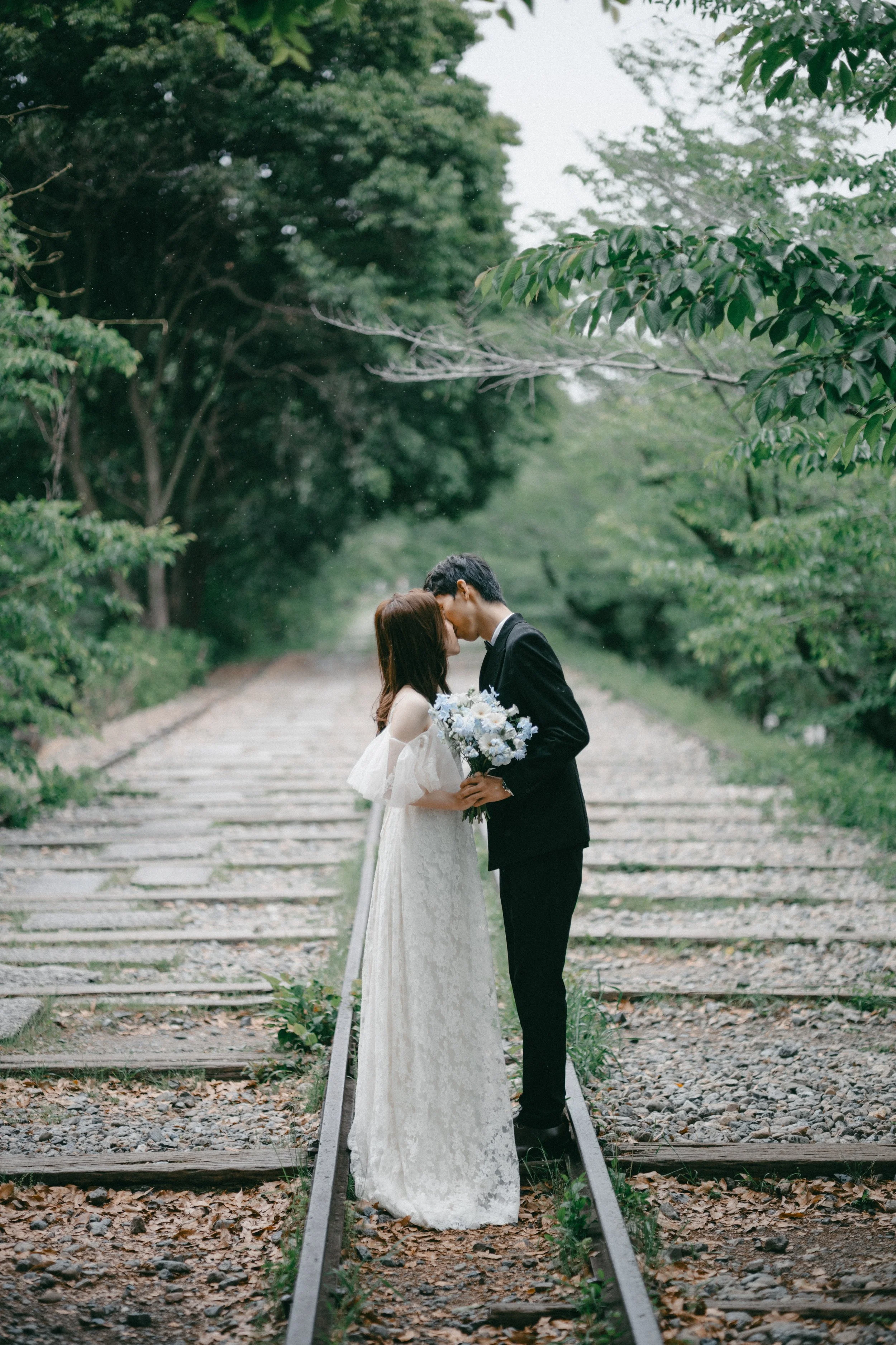 A bride and groom sharing a kiss on a railroad track in a green, wooded outdoor setting, with the bride holding a bouquet of flowers.