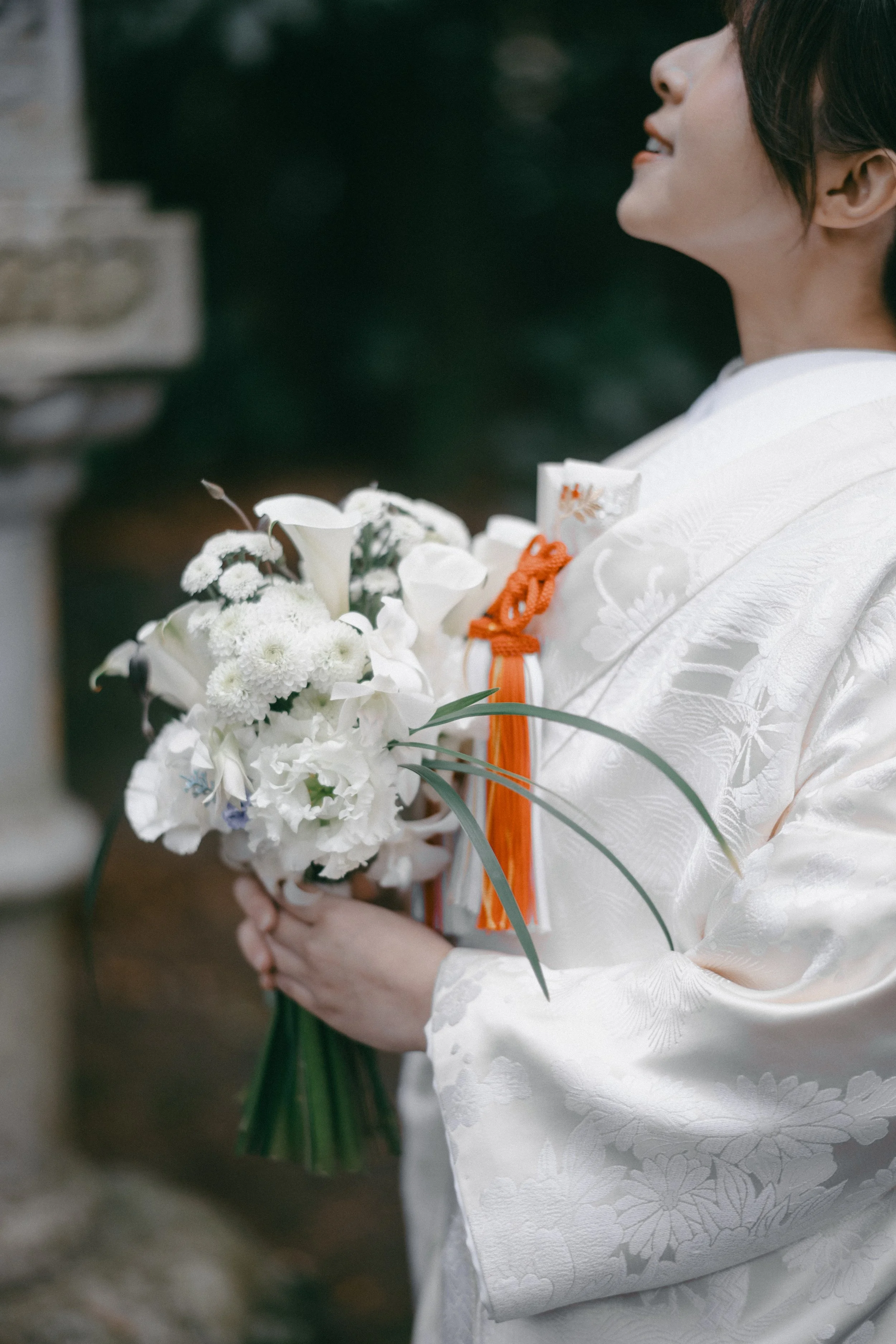 Person holding a bouquet of white flowers and wearing a traditional white kimono with orange accessories, standing outdoors.