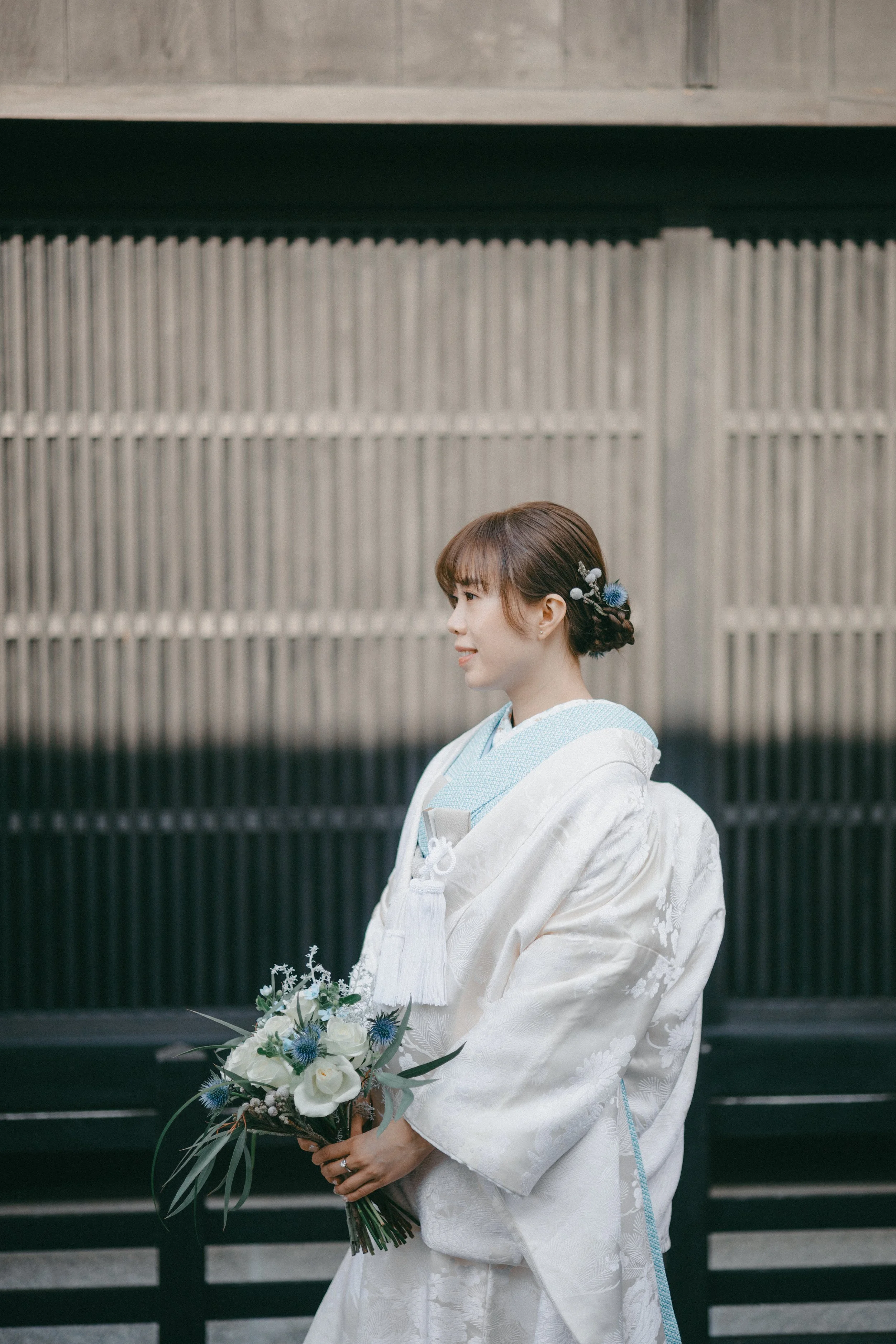 Young woman in traditional Japanese kimono holding a bouquet of white and blue flowers, standing outdoors against a wooden fence background.