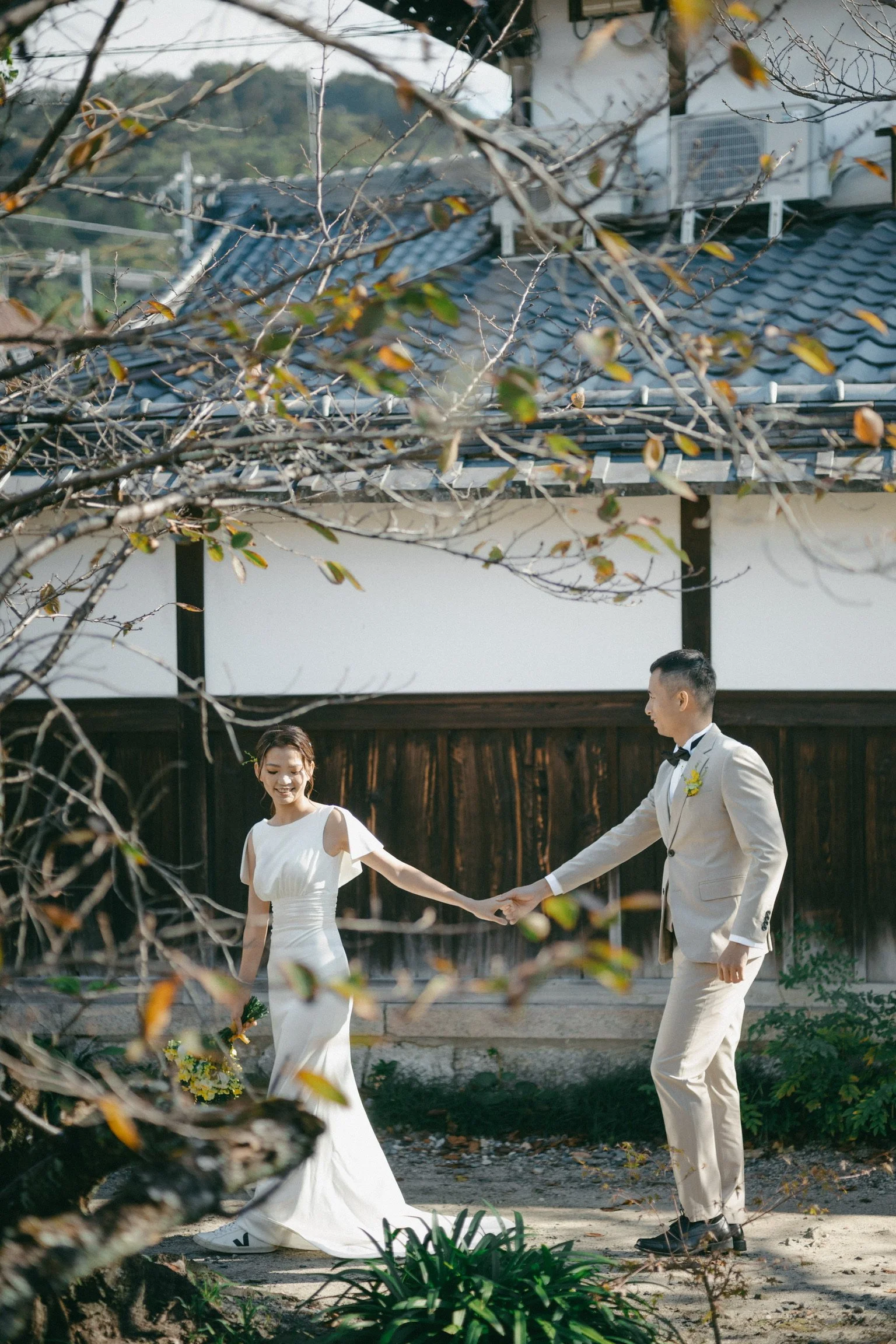A bride and groom holding hands and dancing outdoors during their wedding, with trees and a building in the background.