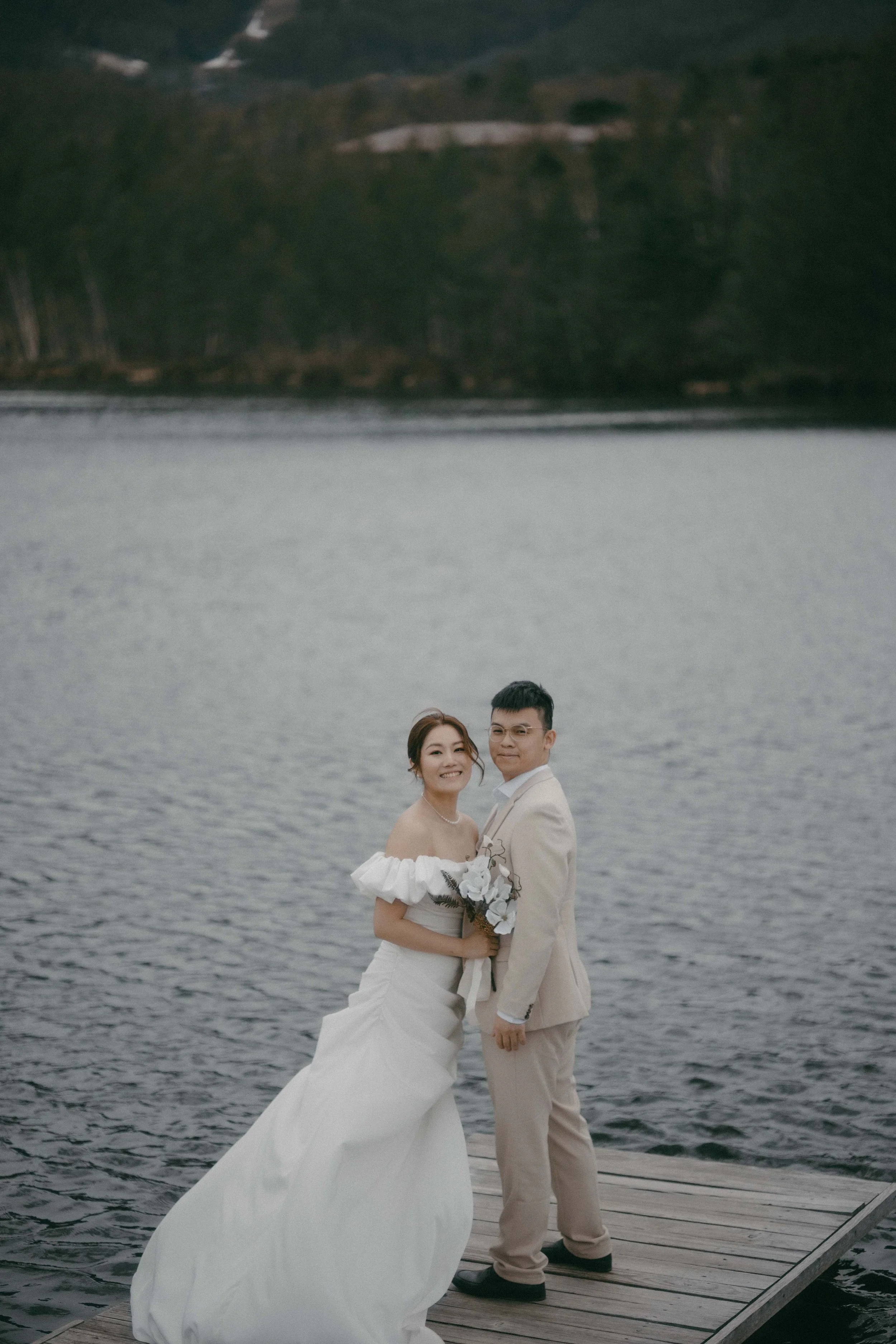 A bride and groom standing on a dock by a lake, with mountains in the background, smiling and posing for a wedding photo.