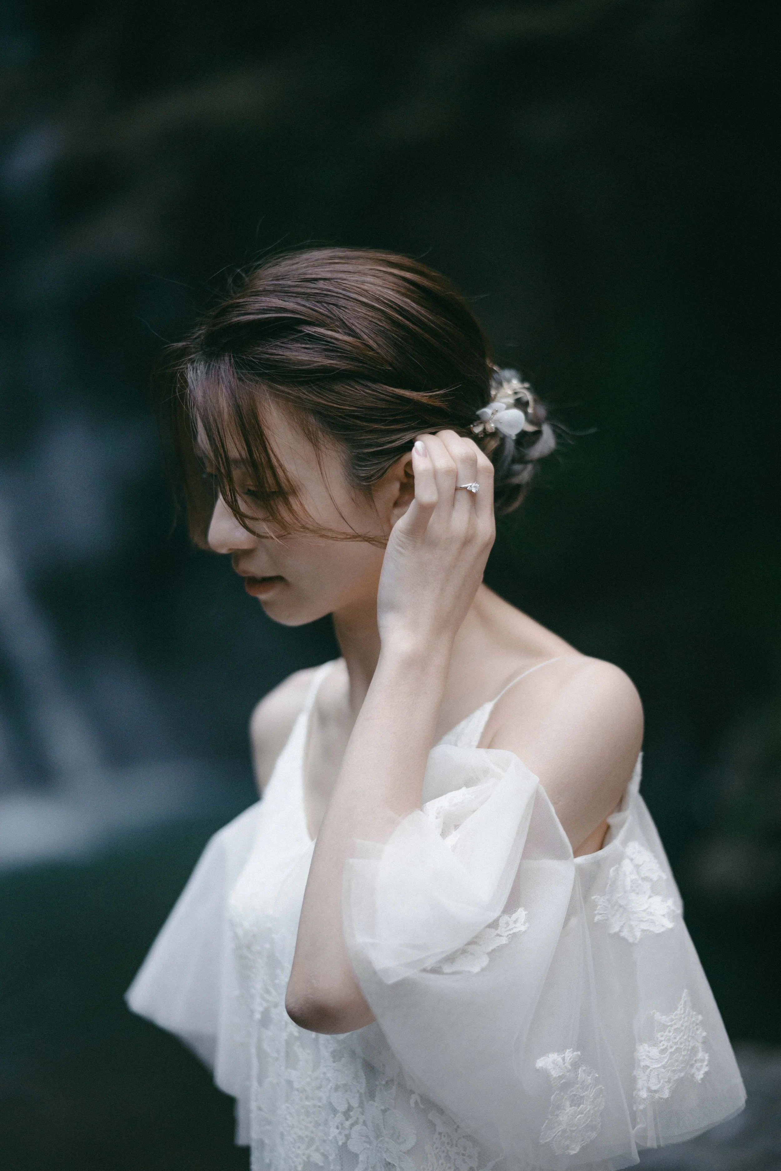 A woman in a white lace dress with puffy sleeves, holding her hair back with her right hand, standing outdoors with a blurred natural background.