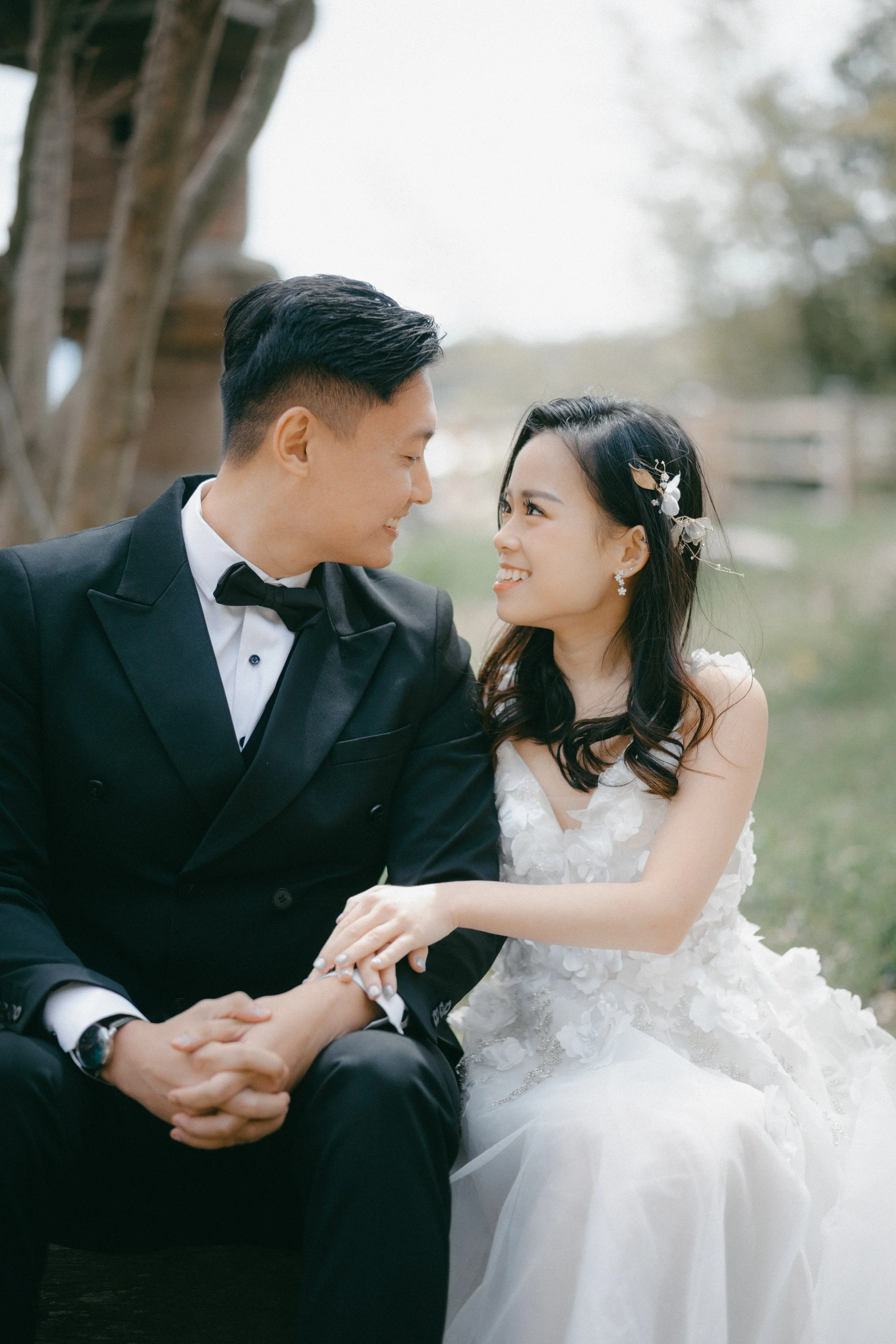 Bride and groom sitting outdoors, smiling at each other, holding hands, in wedding attire.