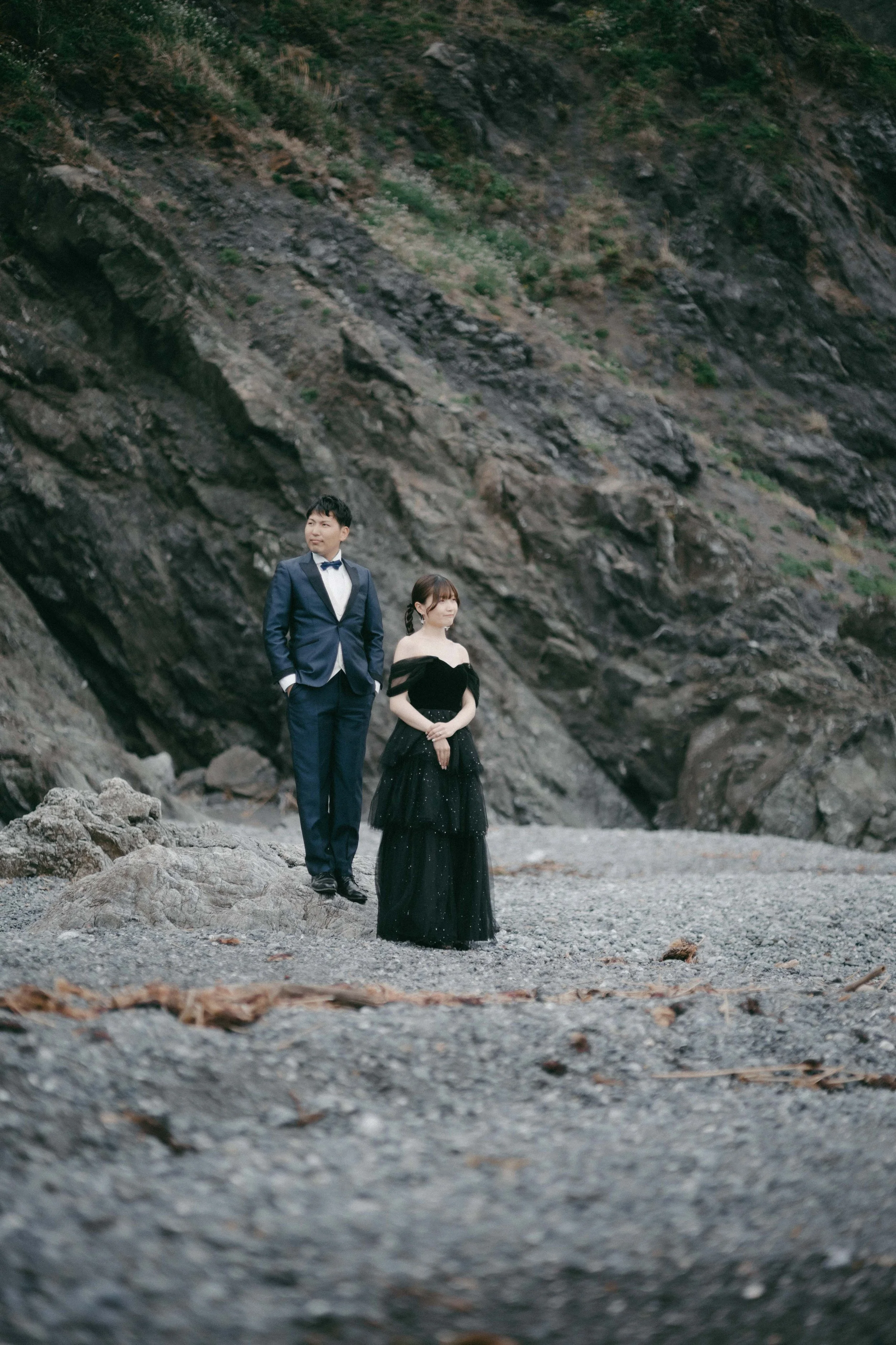 A man in a navy tuxedo and woman in a long black gown standing on a rocky beach against a steep, green hillside.