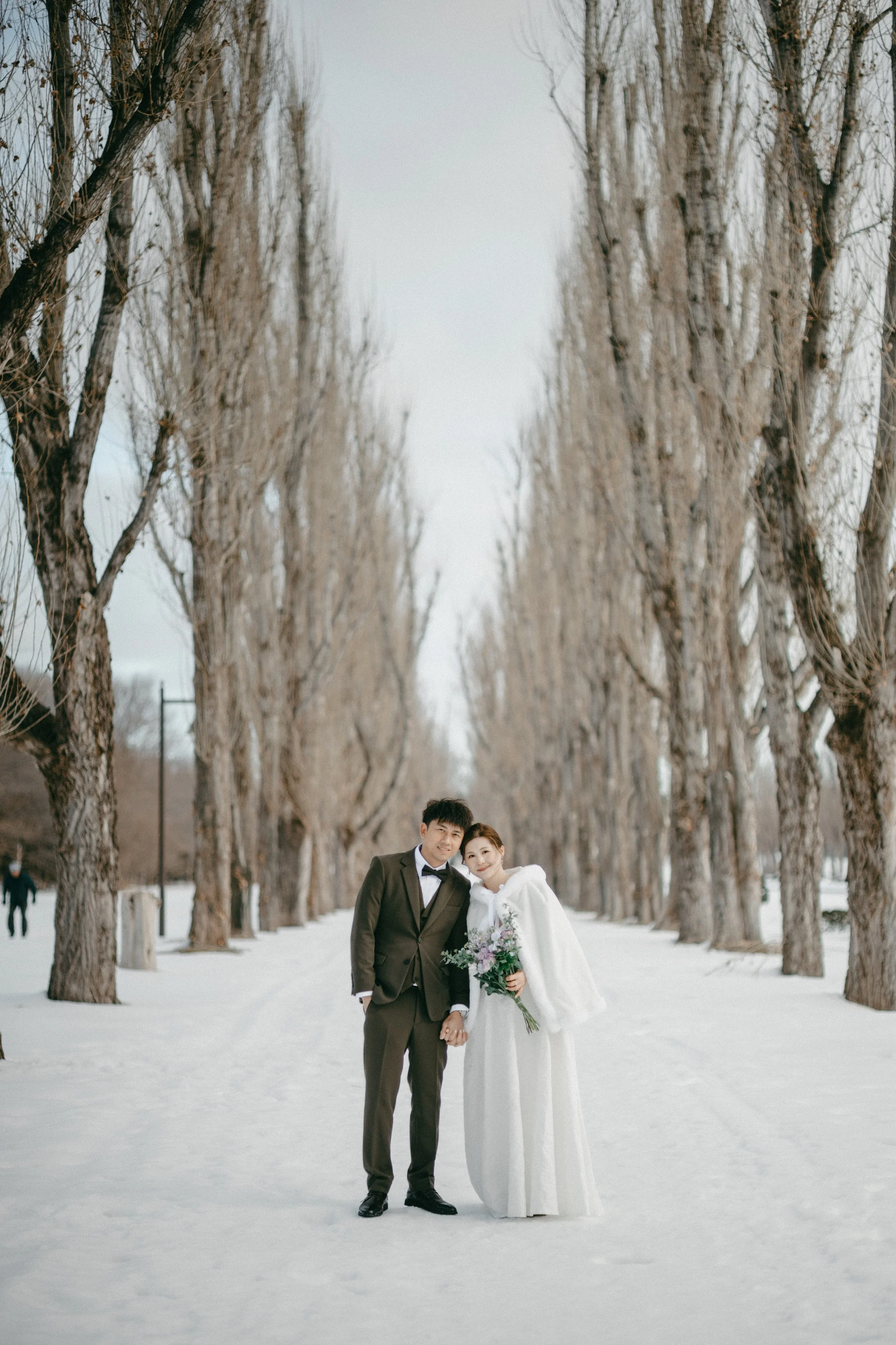 A couple dressed in wedding attire, holding hands and smiling, standing on a snow-covered path lined with tall, leafless trees on a winter day.