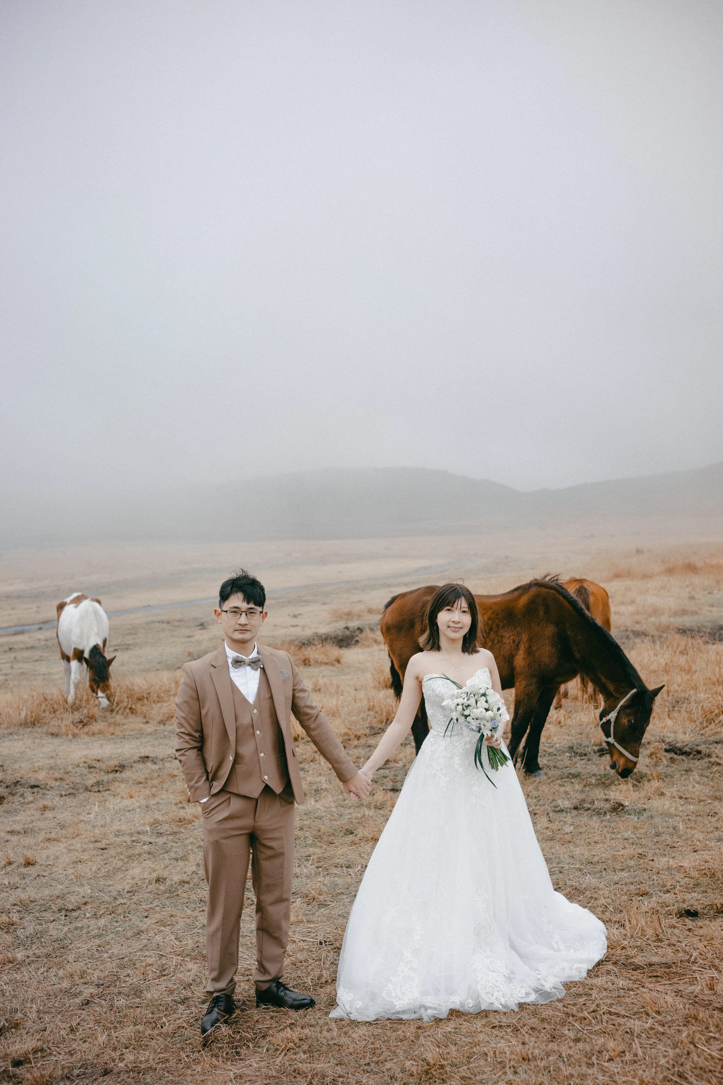 Couple in wedding attire holding hands in a field with horses grazing, overcast sky.