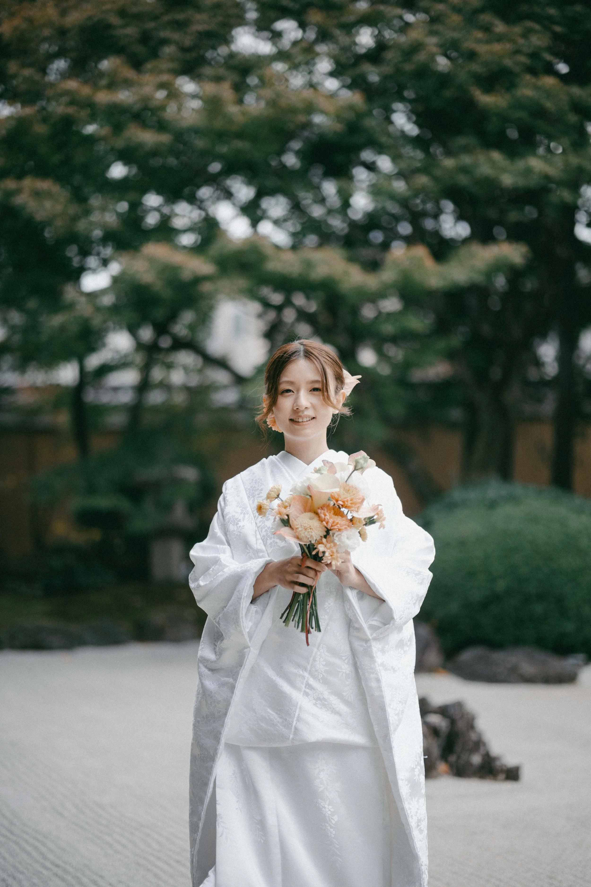 A woman in a traditional white kimono holding a peach-colored bouquet of flowers outdoors in a Japanese garden.