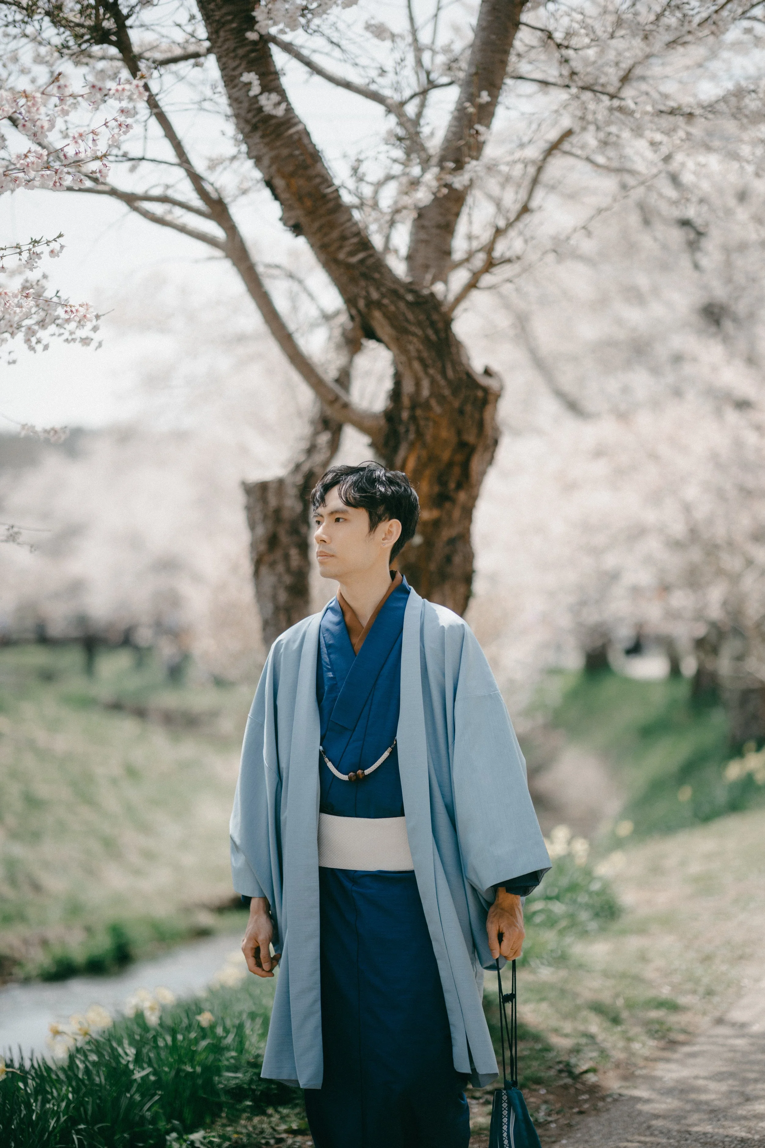 A man dressed in traditional Japanese clothing standing outdoors near blooming cherry blossom trees.