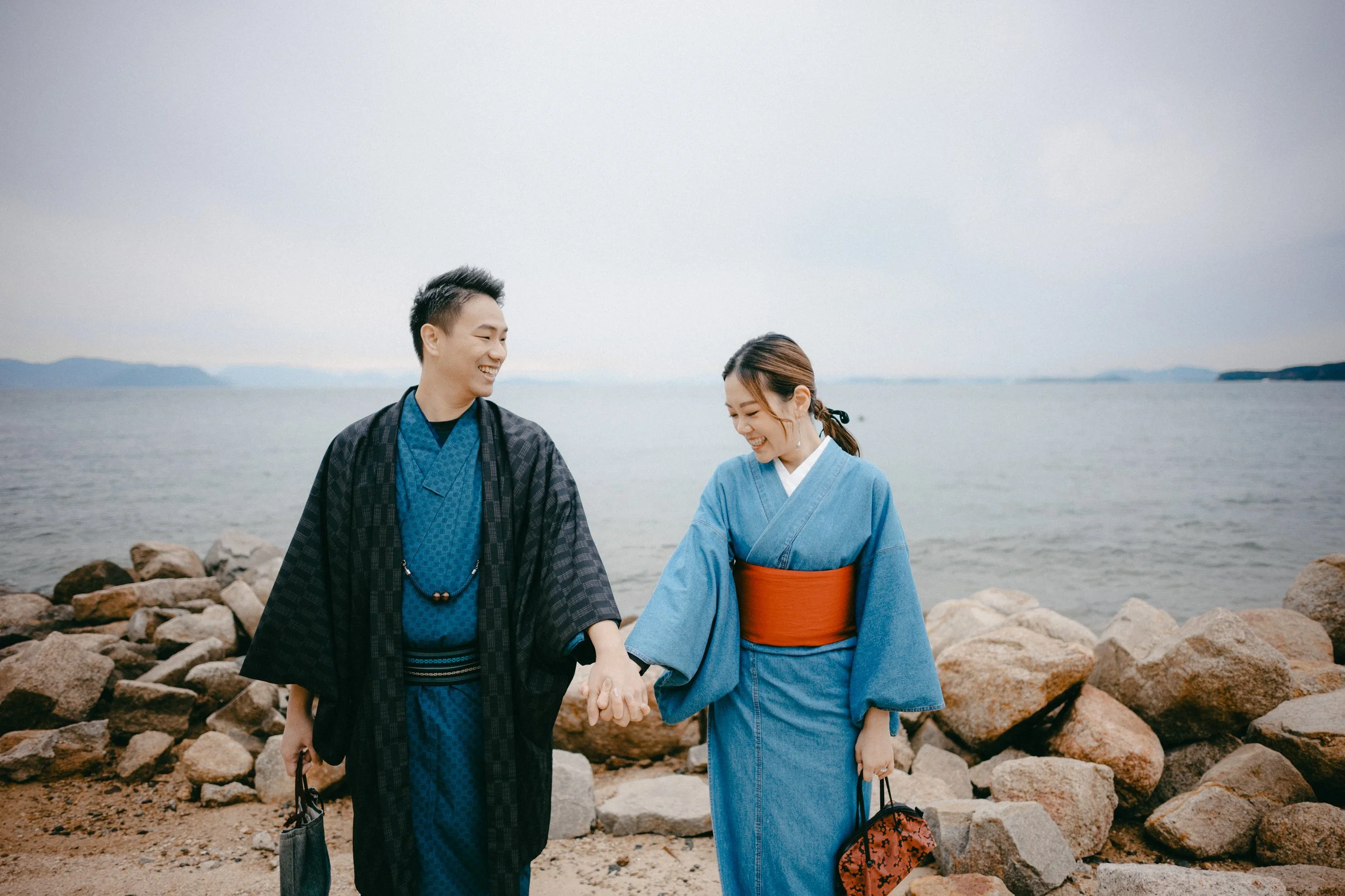 A young couple dressed in traditional Japanese yukatas, holding hands and smiling by the rocky shoreline of a lake or ocean on a cloudy day.