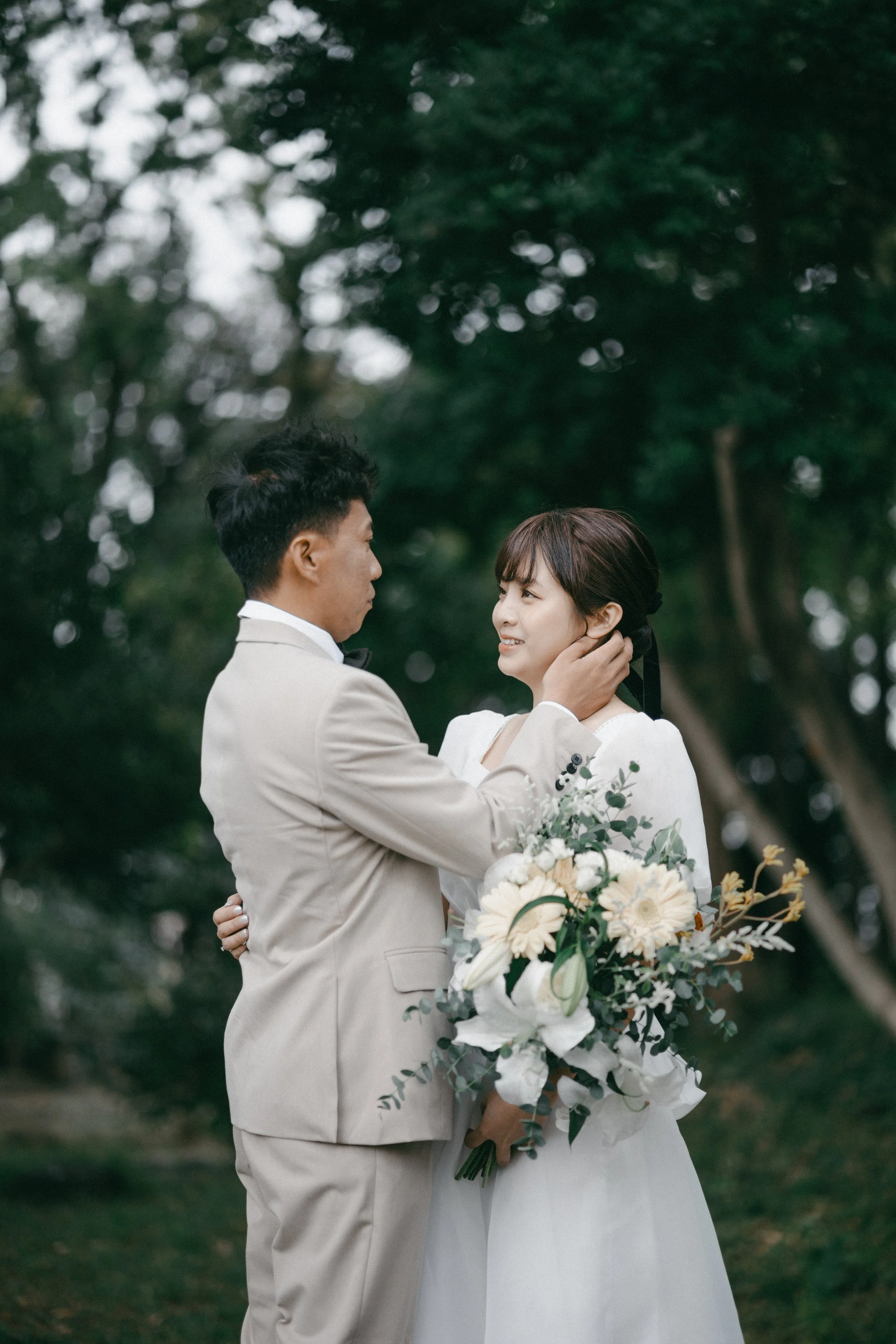 A bride and groom standing outdoors in a natural setting, with the groom touching the bride's face as she holds a bouquet of white and green flowers, sharing an emotional moment.