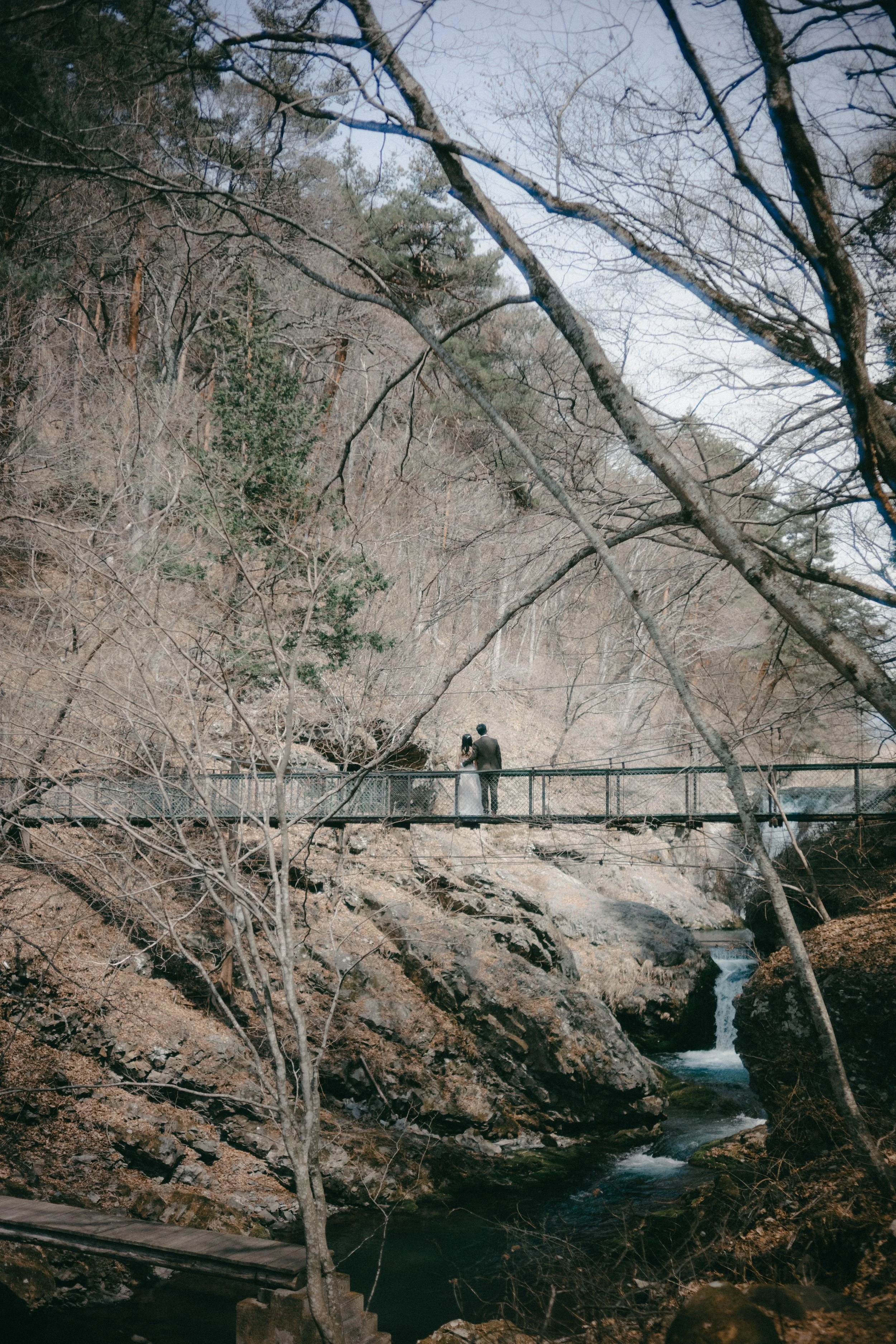 A couple in wedding attire standing on a small bridge over a rocky creek in a wooded area, with leafless trees surrounding them.