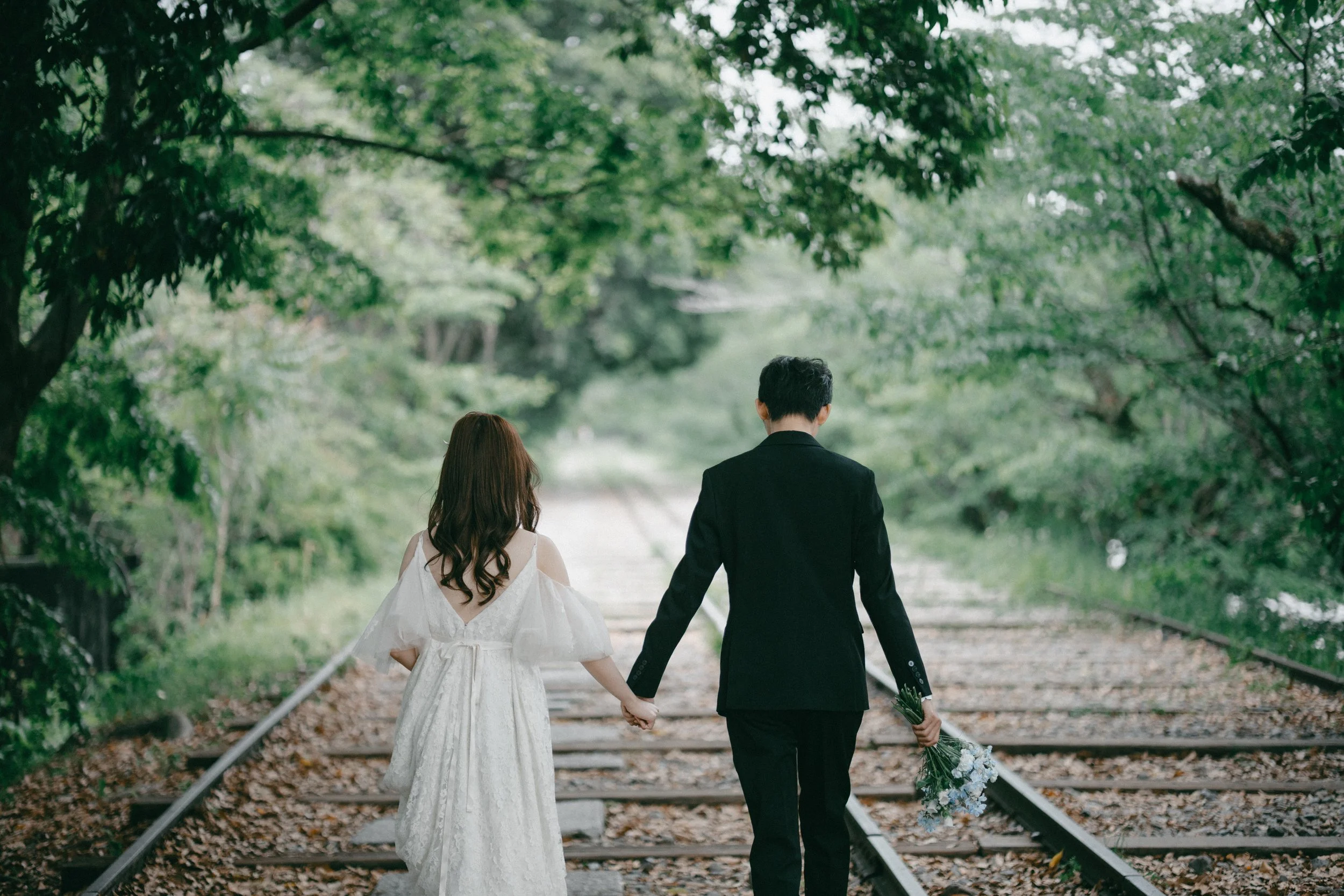 A couple, a bride in a white wedding dress and a groom in a black suit, holding hands and walking along train tracks through a lush, green wooded area. The bride is holding a bouquet of white and blue flowers.