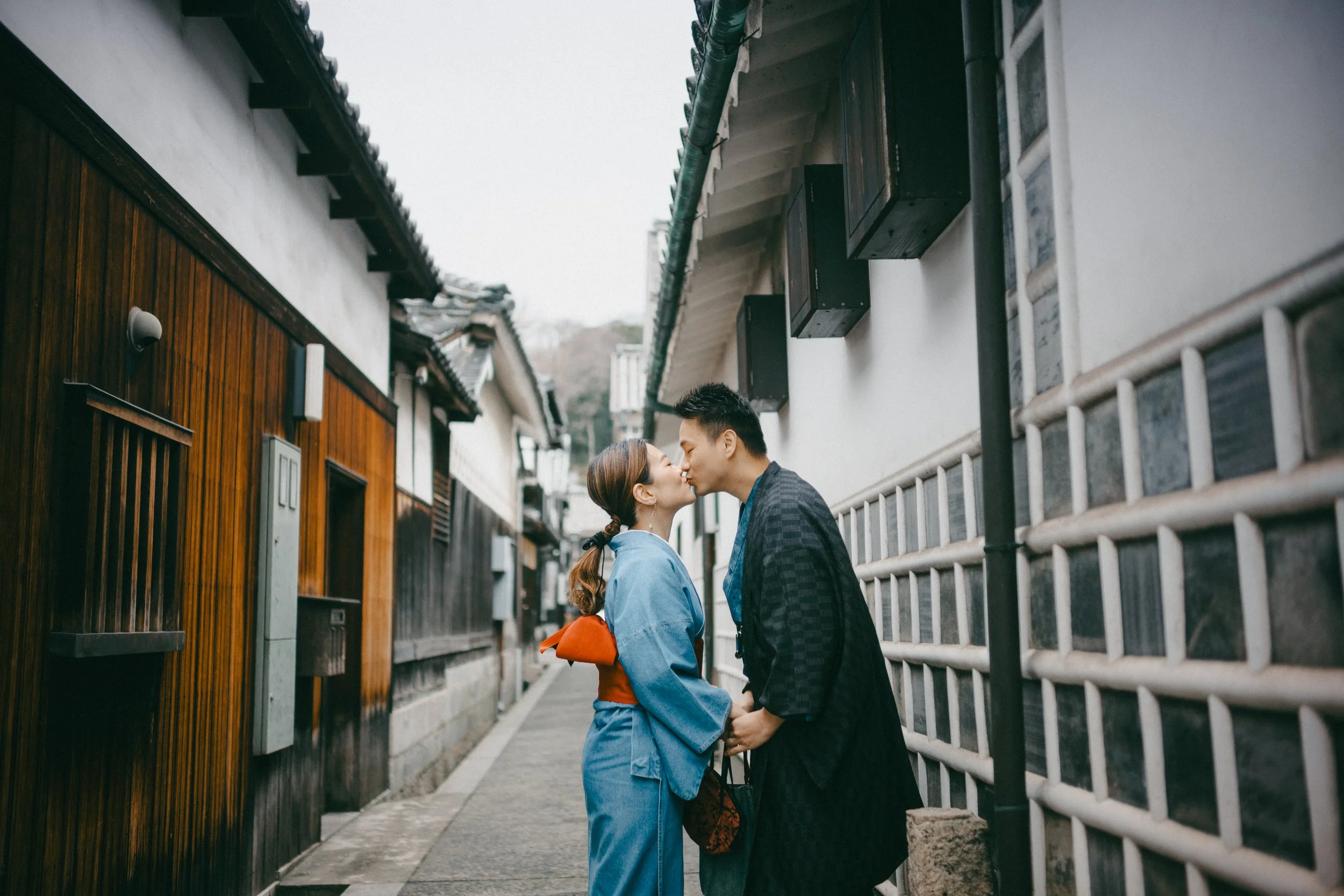 A couple kissing in a narrow alleyway lined with traditional Japanese buildings, with the woman wearing a blue kimono and the man in a dark kimono.