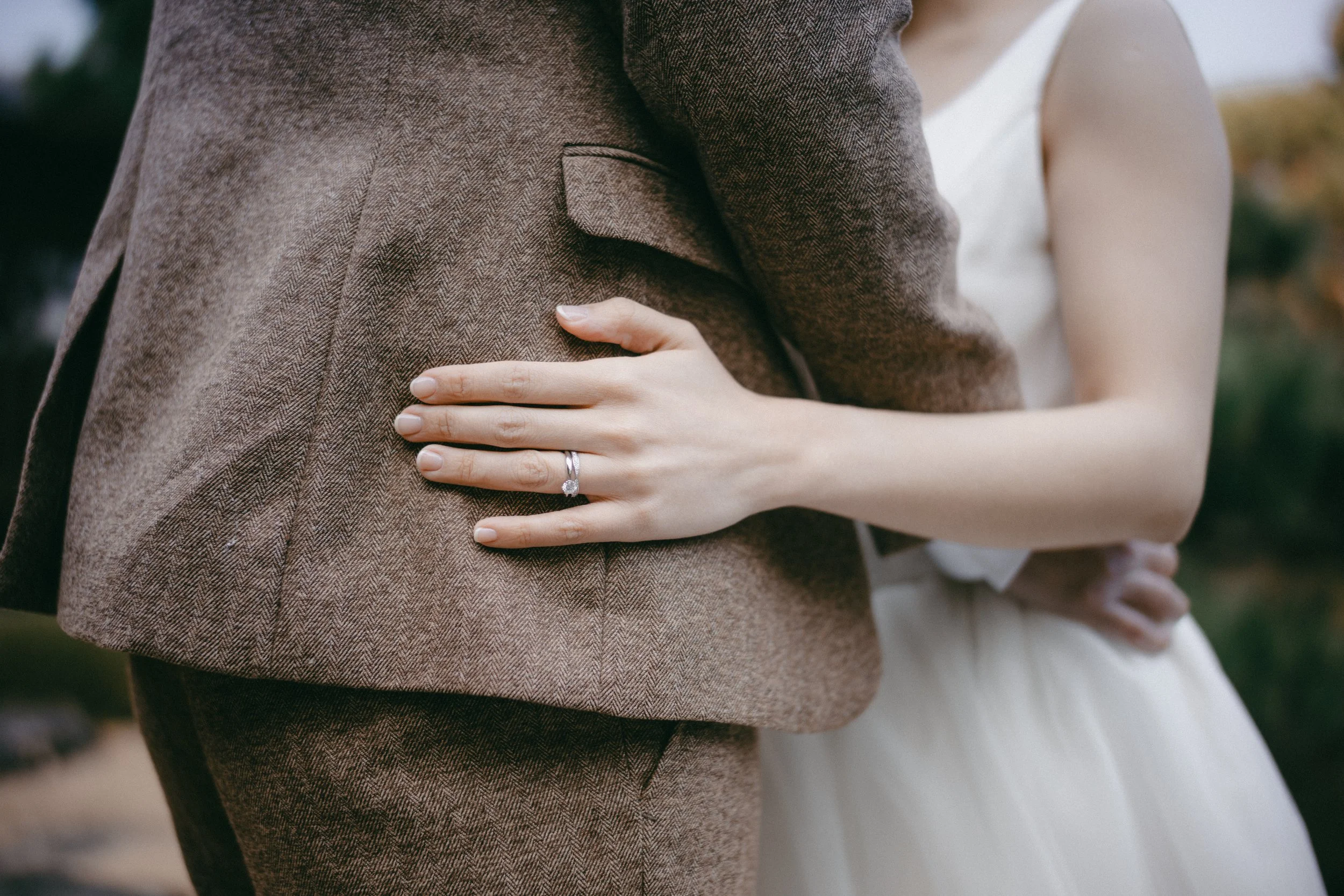 A couple with their arms around each other, the woman wearing a wedding ring, standing outdoors.