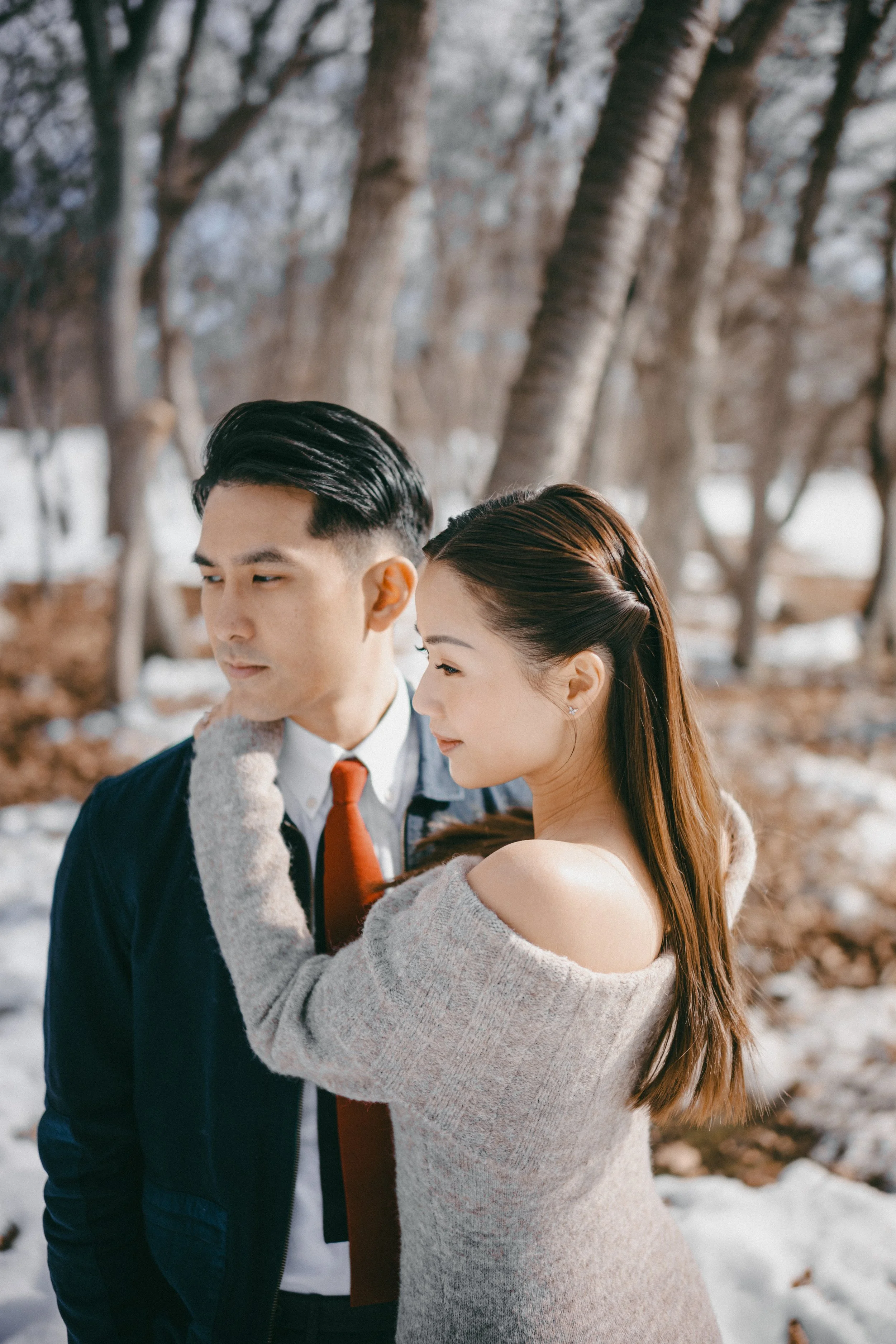 A couple standing close together outdoors in a winter setting with snow on the ground and trees in the background, embracing each other with gentle expressions.