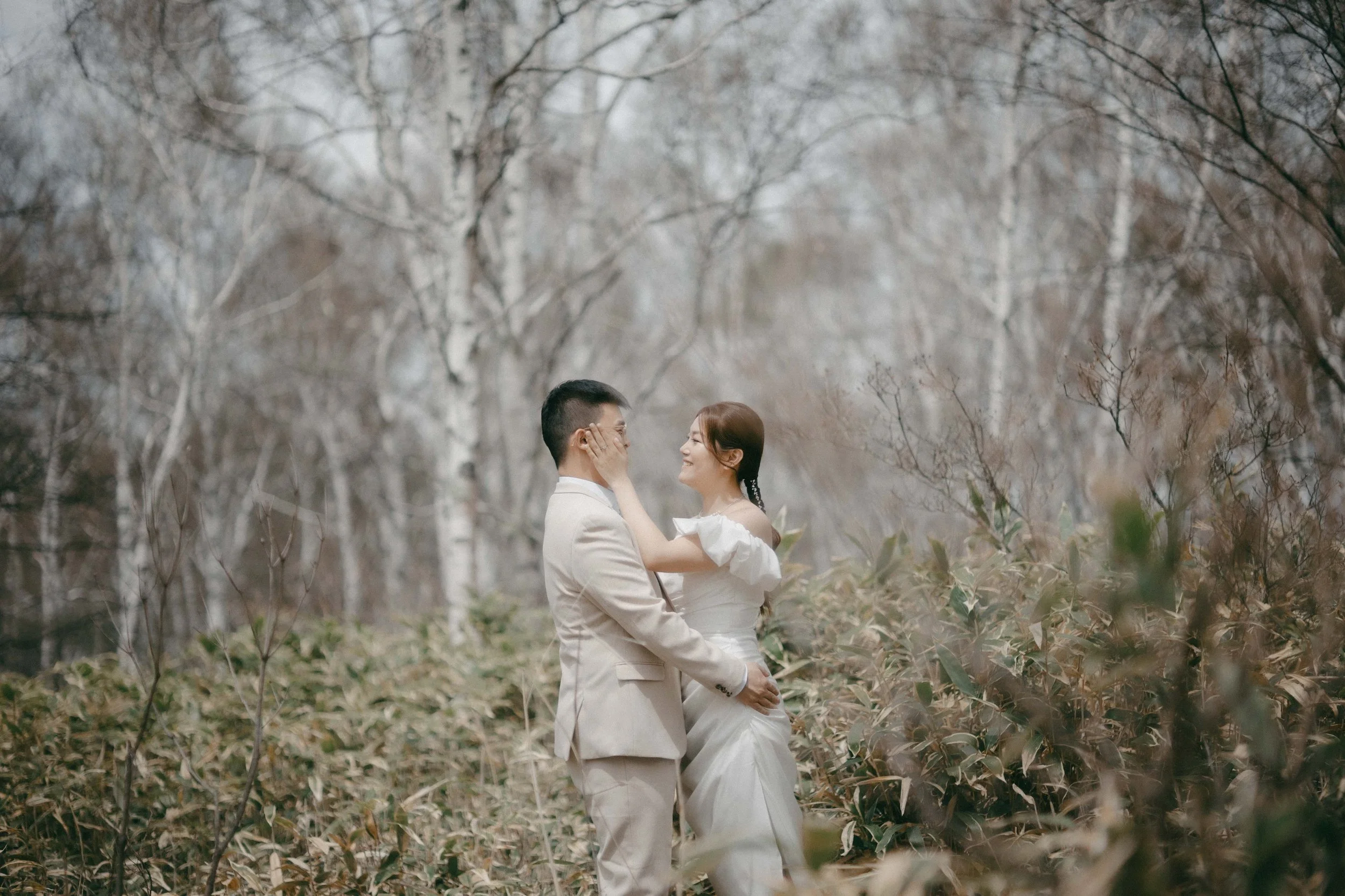 A couple dressed in wedding attire standing in a forest clearing, gazing and smiling at each other with trees and foliage around them.