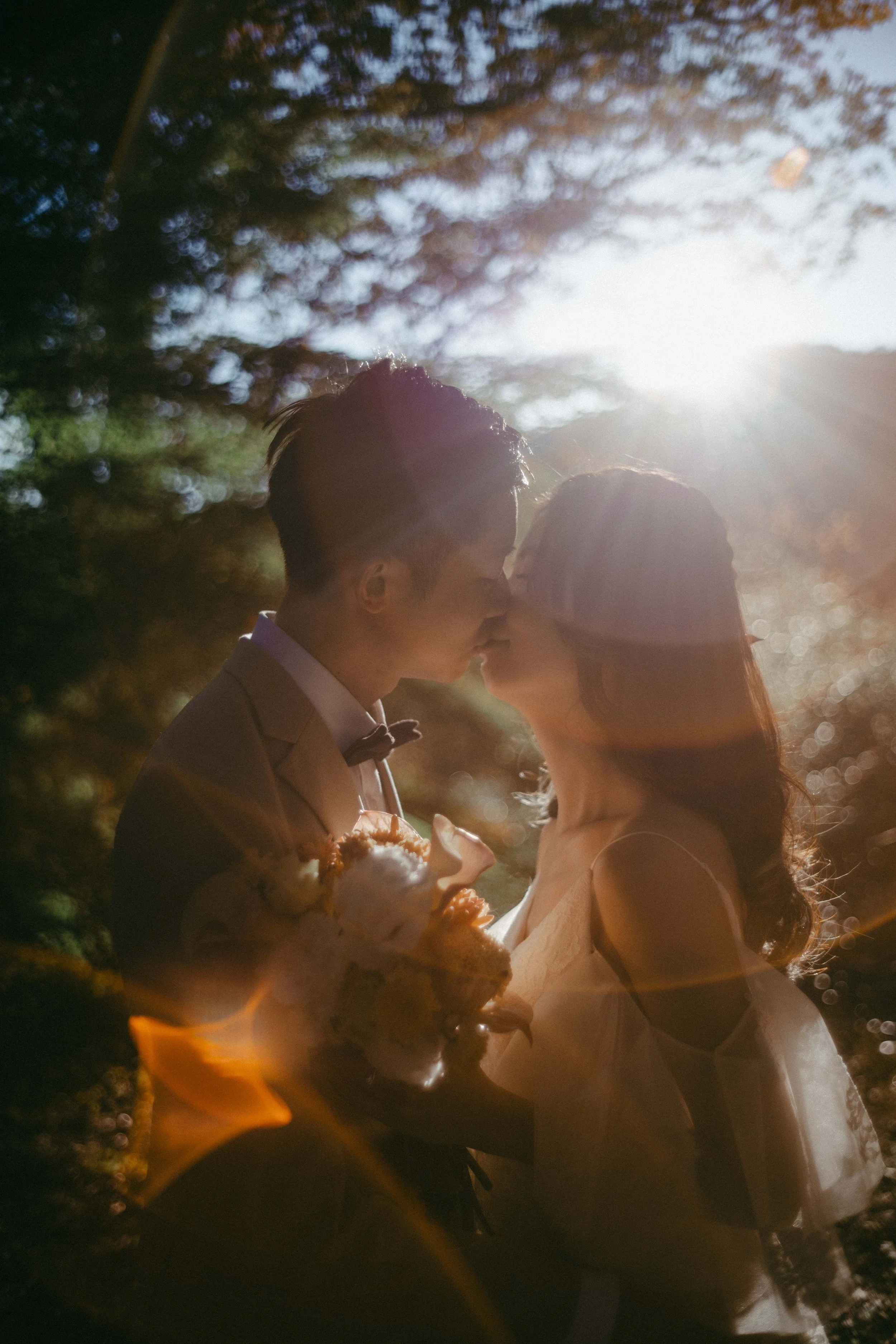 A romantic moment between a newlywed couple kissing outdoors in the sunlight, with the bride holding a bouquet of flowers and both dressed in wedding attire.