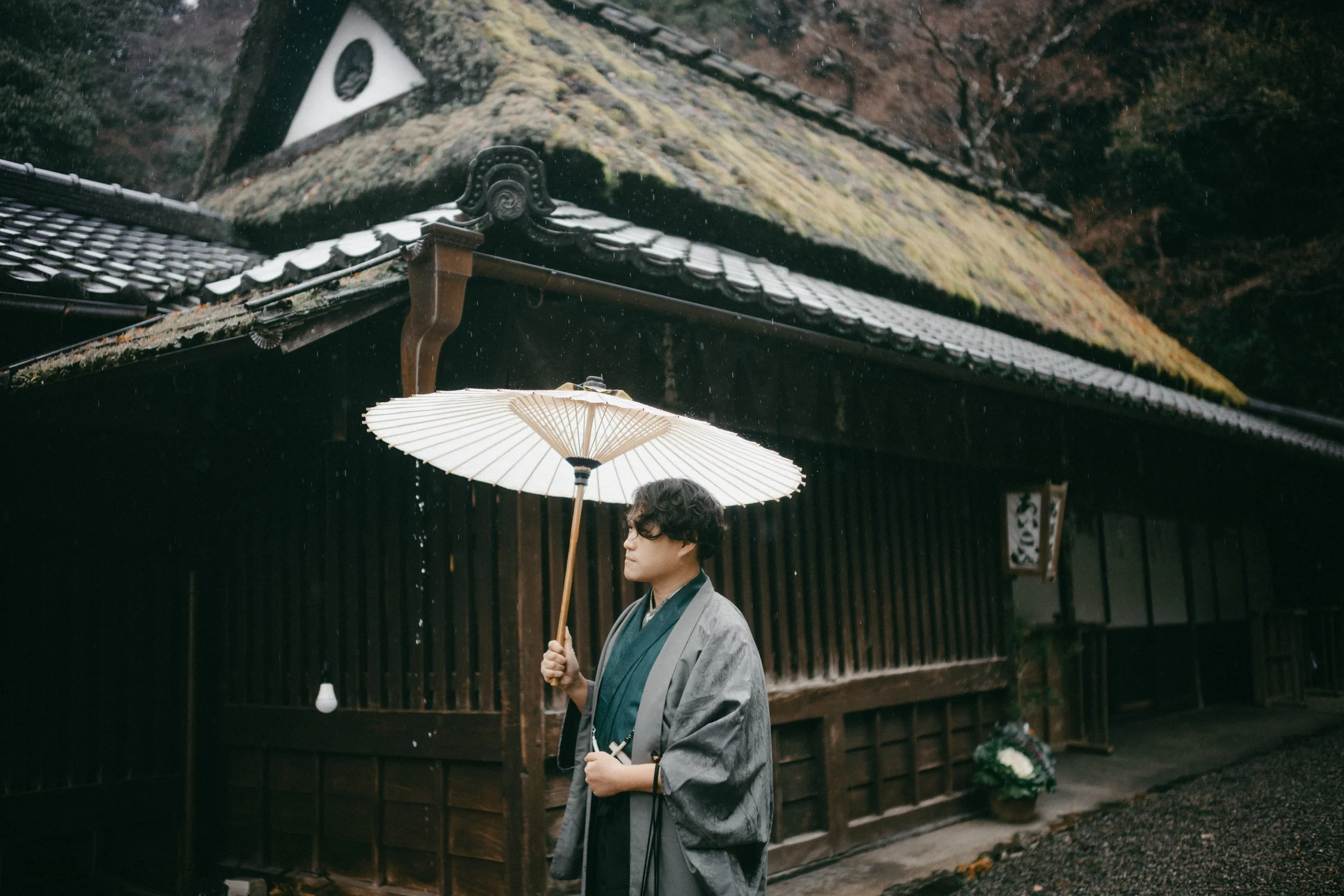 A man dressed in traditional Japanese clothing holding a white paper umbrella standing in front of a traditional wooden building with a thatched roof, surrounded by trees and moss-covered structures.