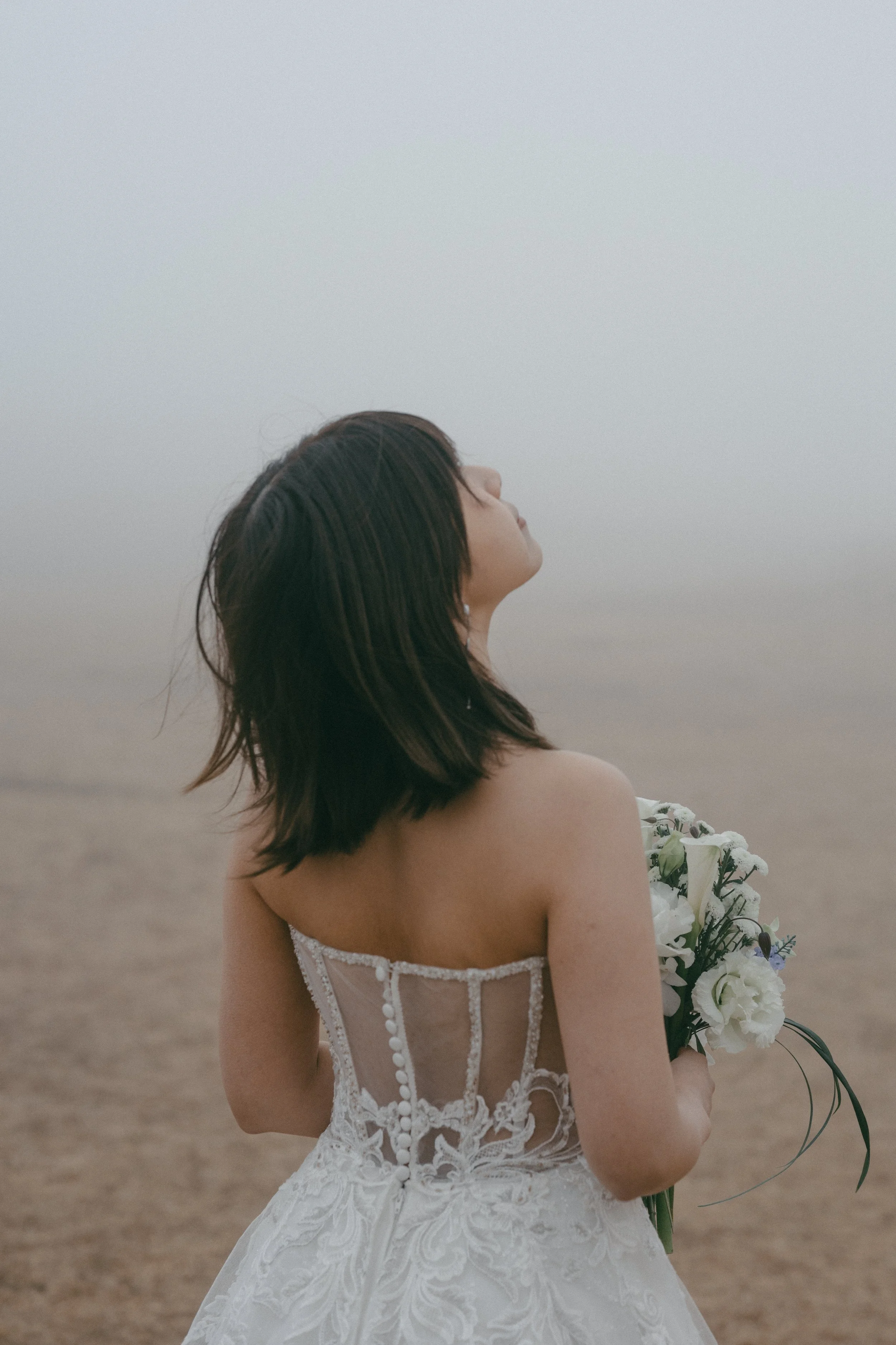 A woman in a wedding dress holding a bouquet of flowers, standing outdoors on a foggy day with her eyes closed.