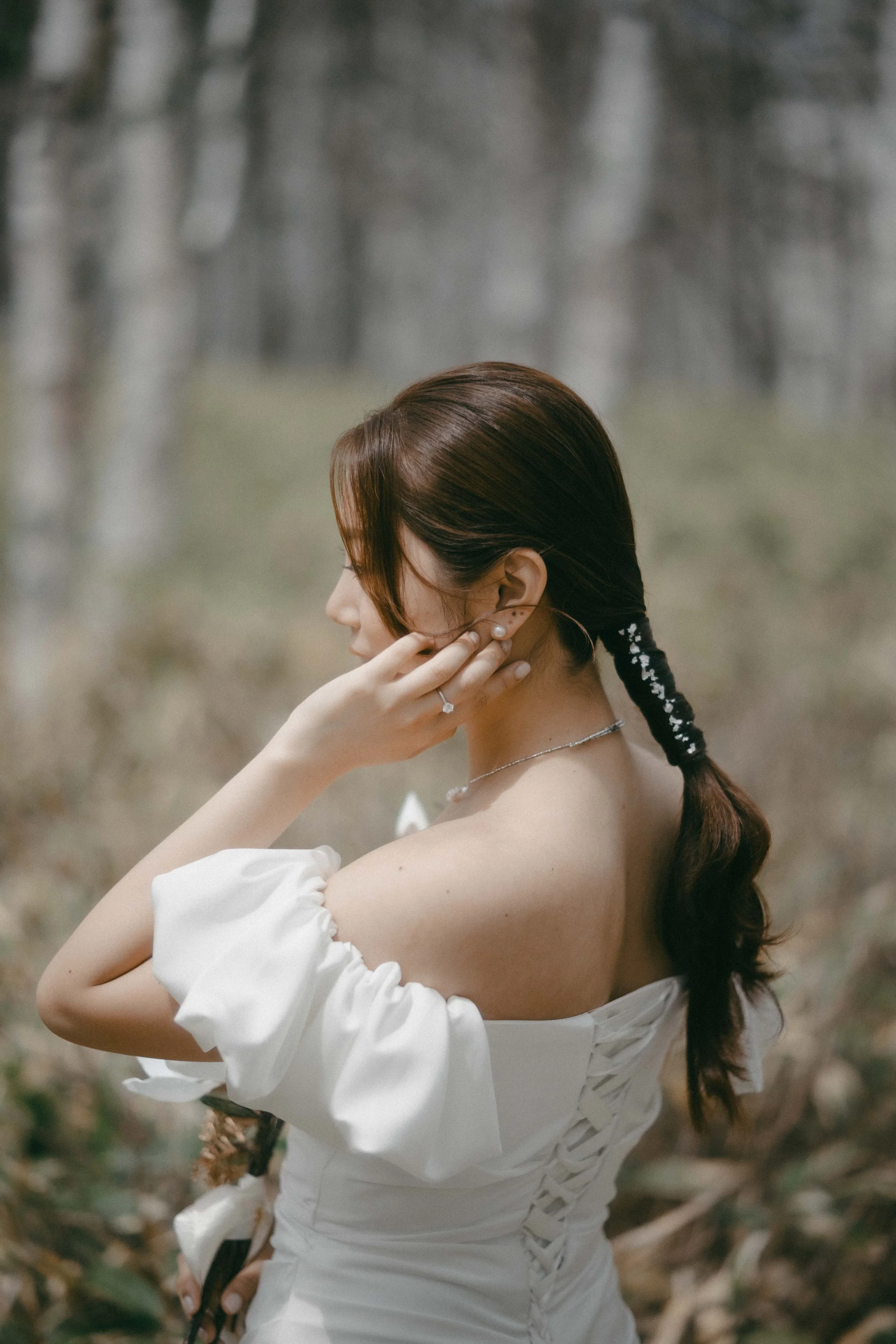 A woman in a white dress with puffed sleeves, standing outdoors in a natural setting with blurred trees in the background. She has brown hair styled in a braid and is wearing jewelry, including earrings, a necklace, and a ring. She is touching her ea