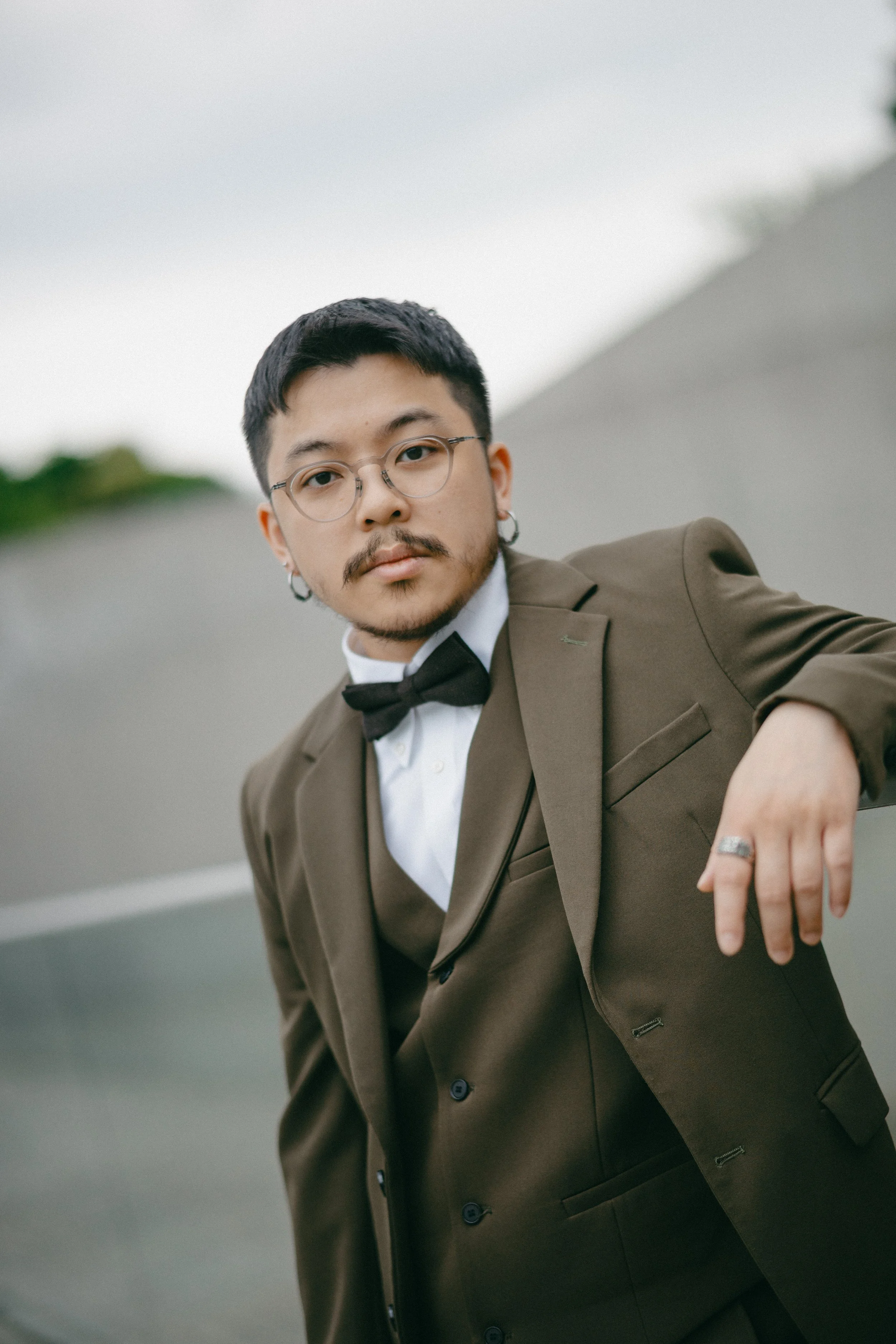 A young man with glasses, earrings, and a mustache wearing a brown suit with a white shirt and black bow tie, standing outdoors on a road with a blurred background.