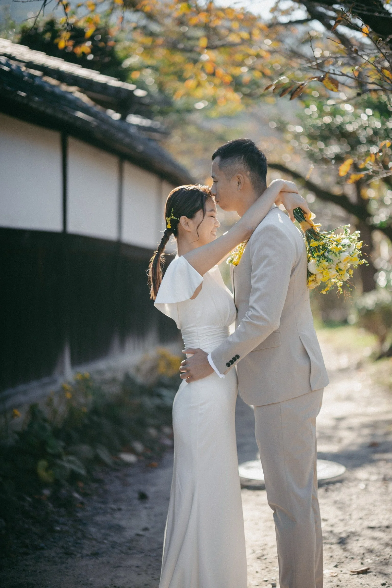A bride and groom sharing a romantic moment outdoors, with the groom kissing the bride's forehead and the bride holding a bouquet of yellow flowers. They are dressed in wedding attire, standing on a path with autumn leaves around them.