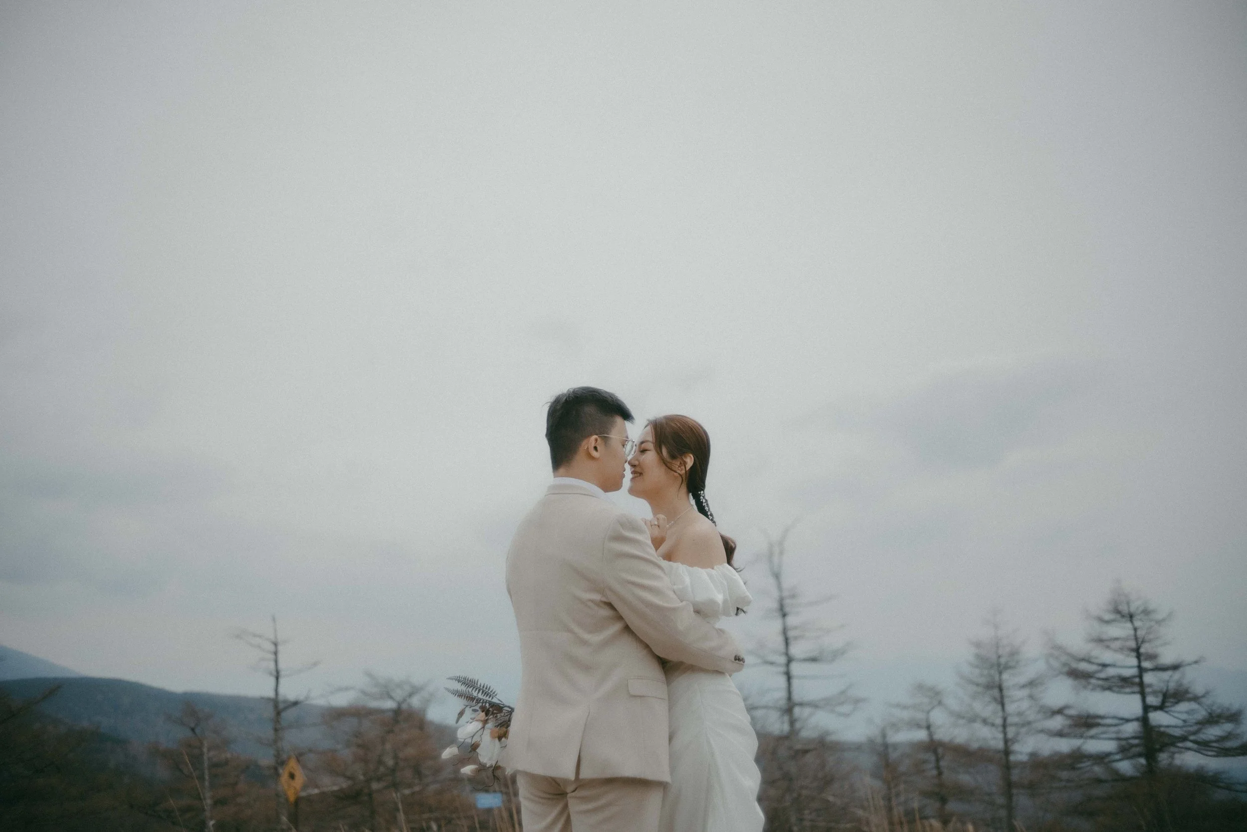 A couple in wedding attire embracing outdoors on a cloudy day with a mountain and barren trees in the background.