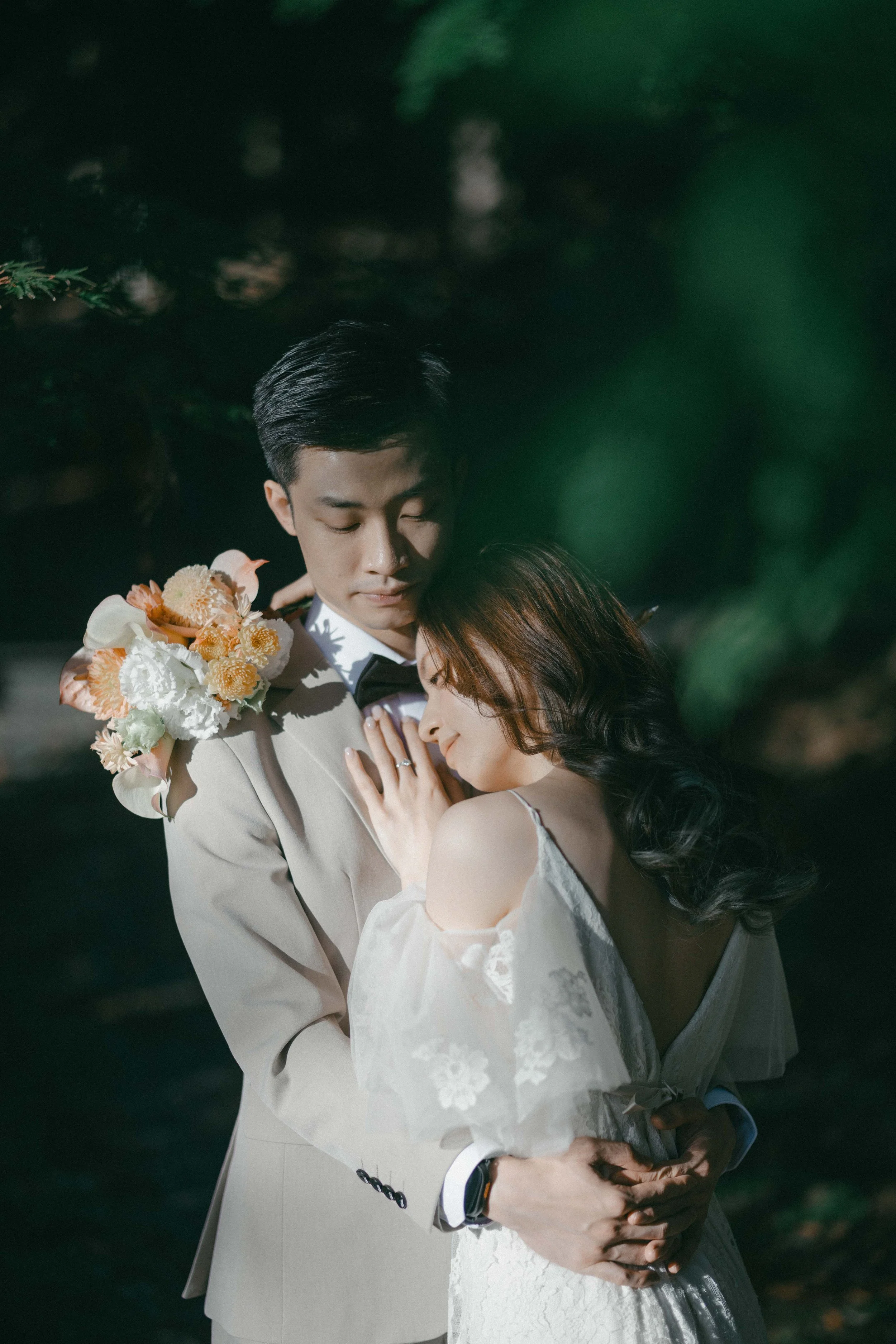 A couple in wedding attire embracing outdoors with eyes closed, surrounded by greenery.