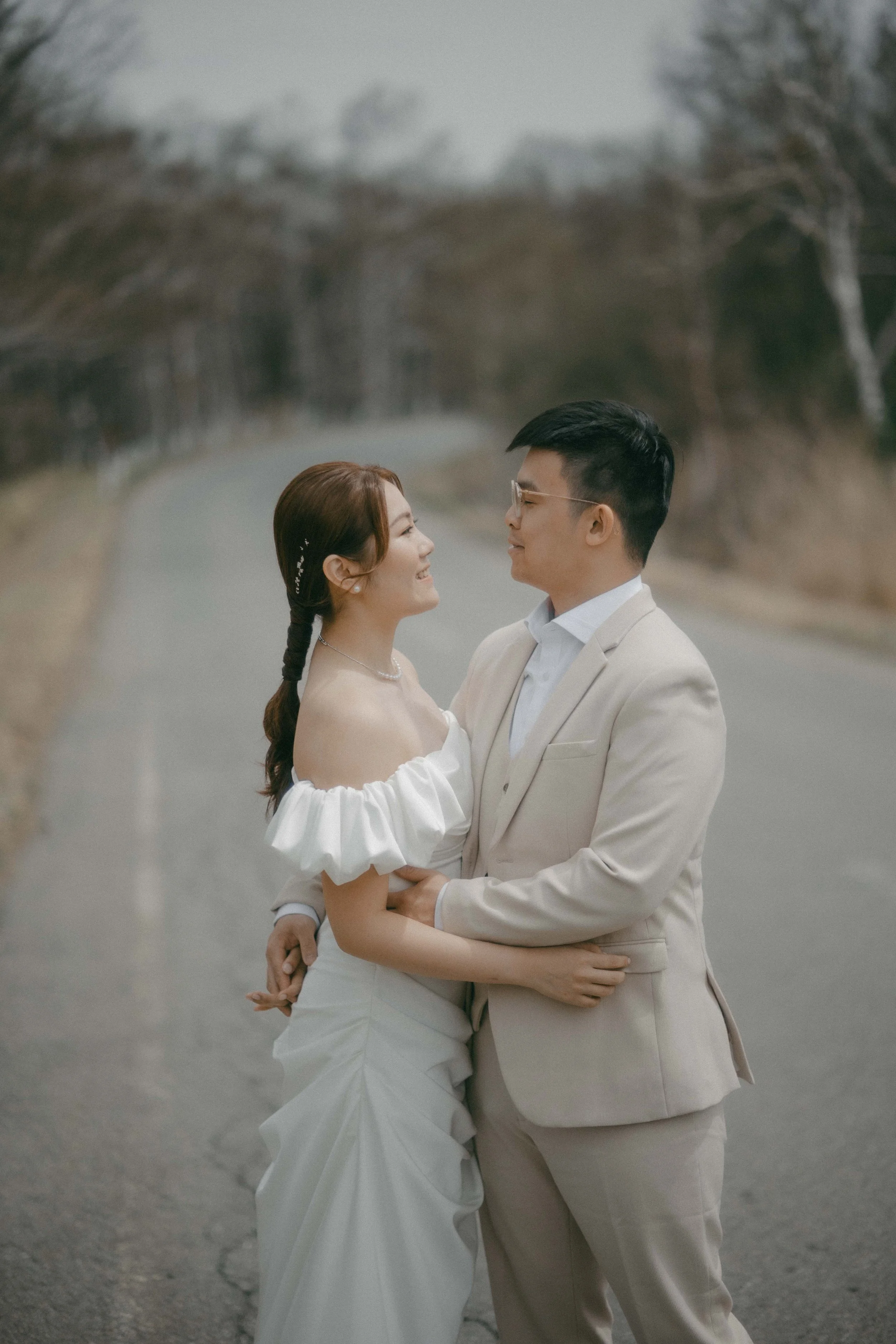A bride and groom standing on a deserted road, smiling at each other, in a countryside setting with leafless trees.