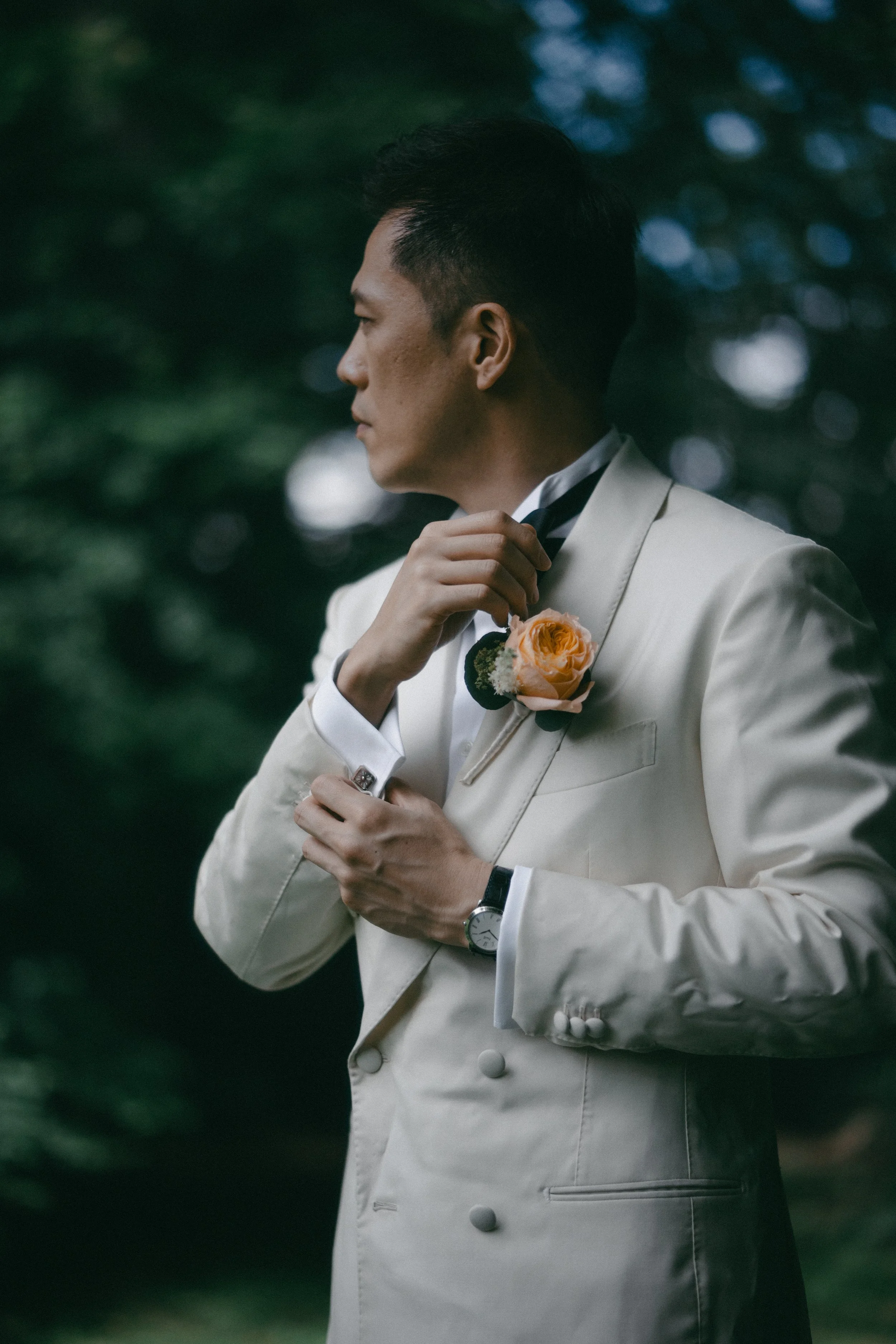 A man dressed in a white tuxedo with a boutonnière, adjusting his bowtie outdoors, with a blurred green leafy background.