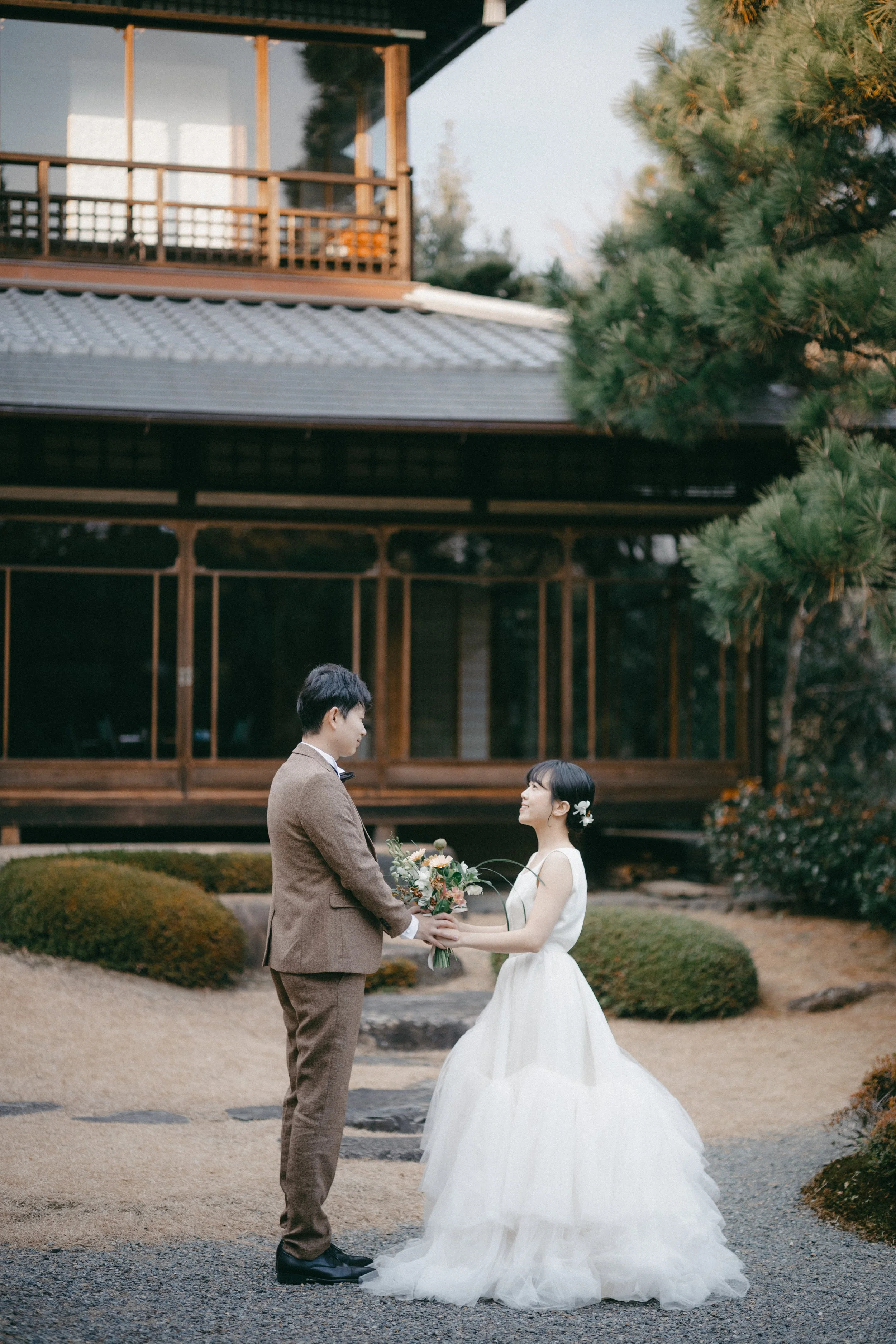 A bride and groom standing face-to-face outdoors, holding flowers, on a gravel path with garden bushes and a traditional Japanese-style building in the background.